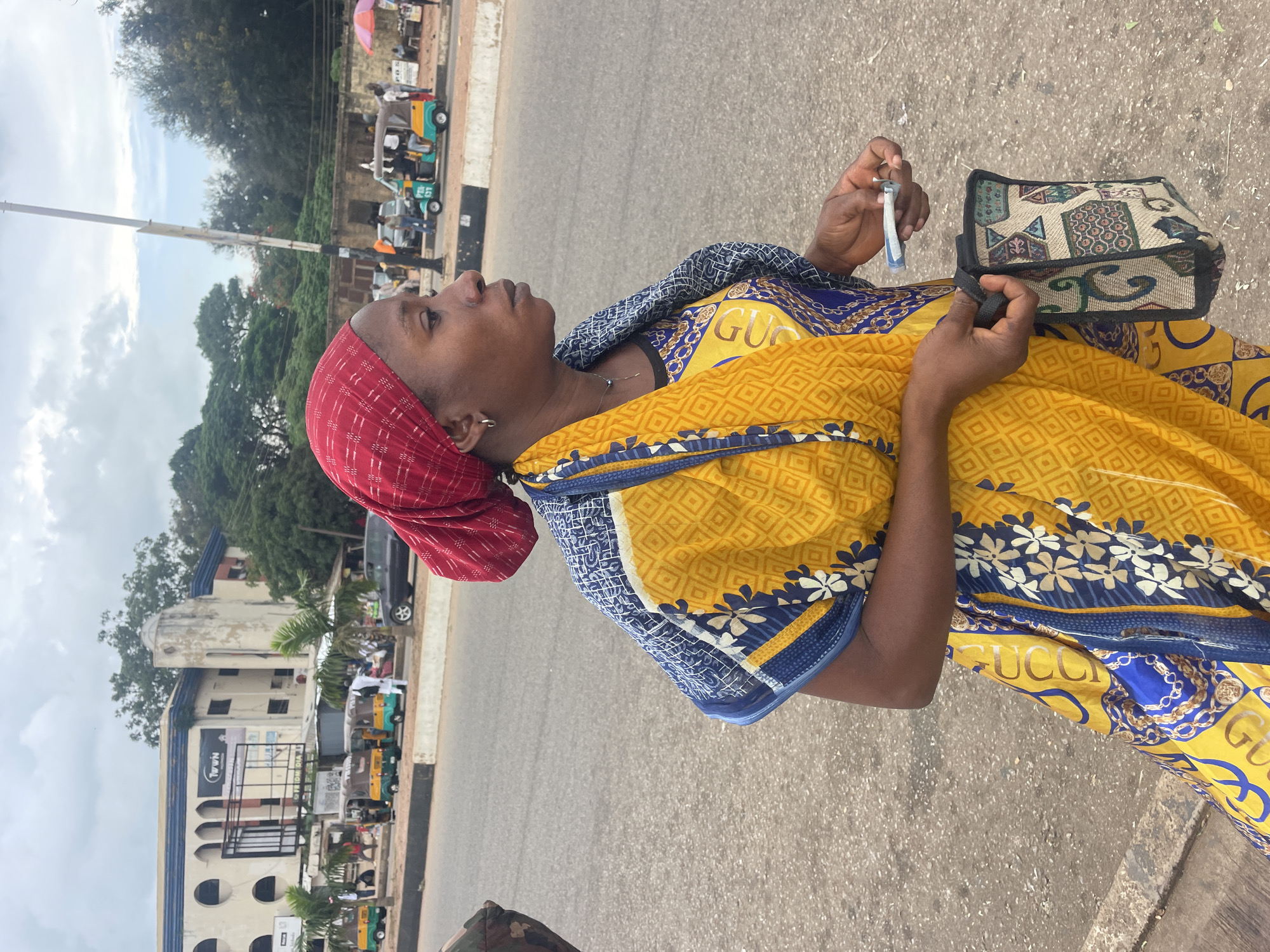 A woman wearing a red headscarf and a yellow and blue patterned dress stands in the street in Barakin Lerawi, Nigeria. She is holding a small handheld device and a patterned bag. In the background, there are several auto-rickshaws, a building with an arched entrance, trees, and a cloudy sky. The street is paved with asphalt.