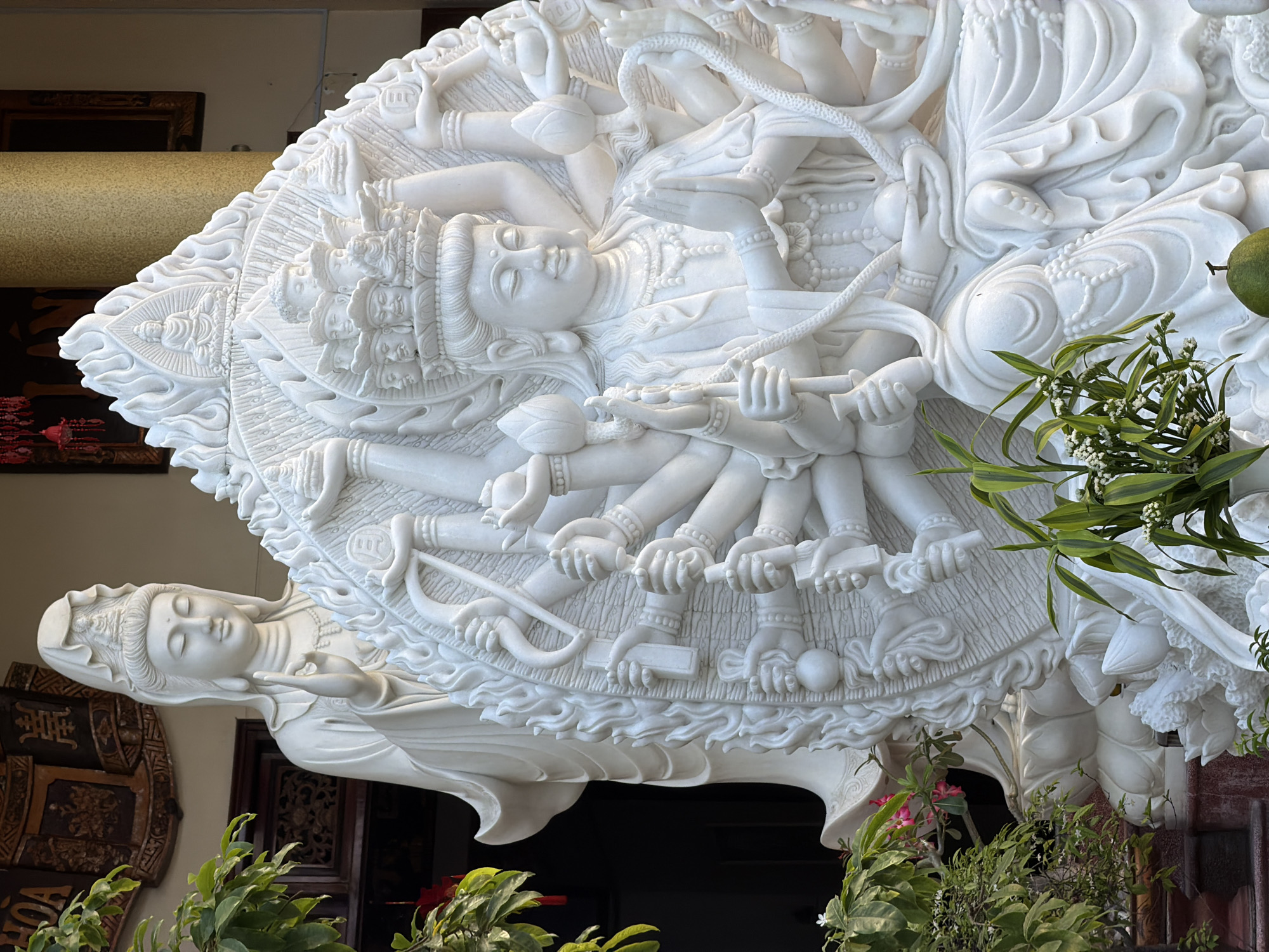 A detailed, ornate, white statue of a deity with multiple arms and serene faces is the focal point of the image. The statue is positioned in a way that suggests it is part of a larger display, possibly in a temple or cultural center. The statue features intricate carvings and a smooth, polished surface. Below the statue, lush greenery and potted plants add a natural element to the scene. The location is Tay Nha Trang Ward, Vietnam. The environment is indicative of a religious or cultural setting.