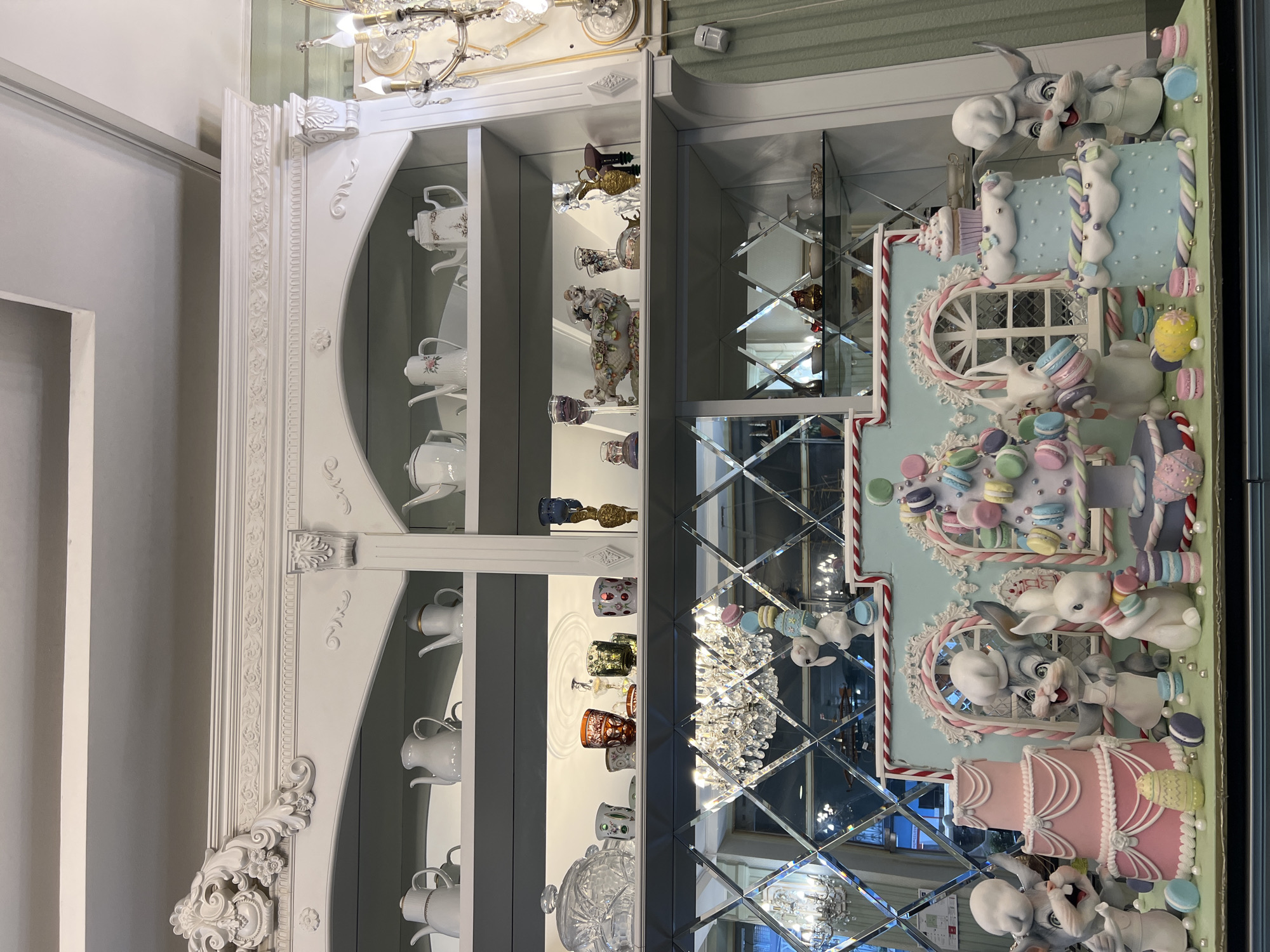 The image captures a display inside a store, likely located in the city of Tbilisi, Georgia. On the left is a white, ornate display cabinet filled with teacups, teapots, and decorative glassware. To the right of the cabinet, attached to the wall, is a decorative display of cartoonish rabbits and desserts such as cakes and macarons. A mirror with diamond-shaped beveled edges is behind the decorations, reflecting the interior of the store.