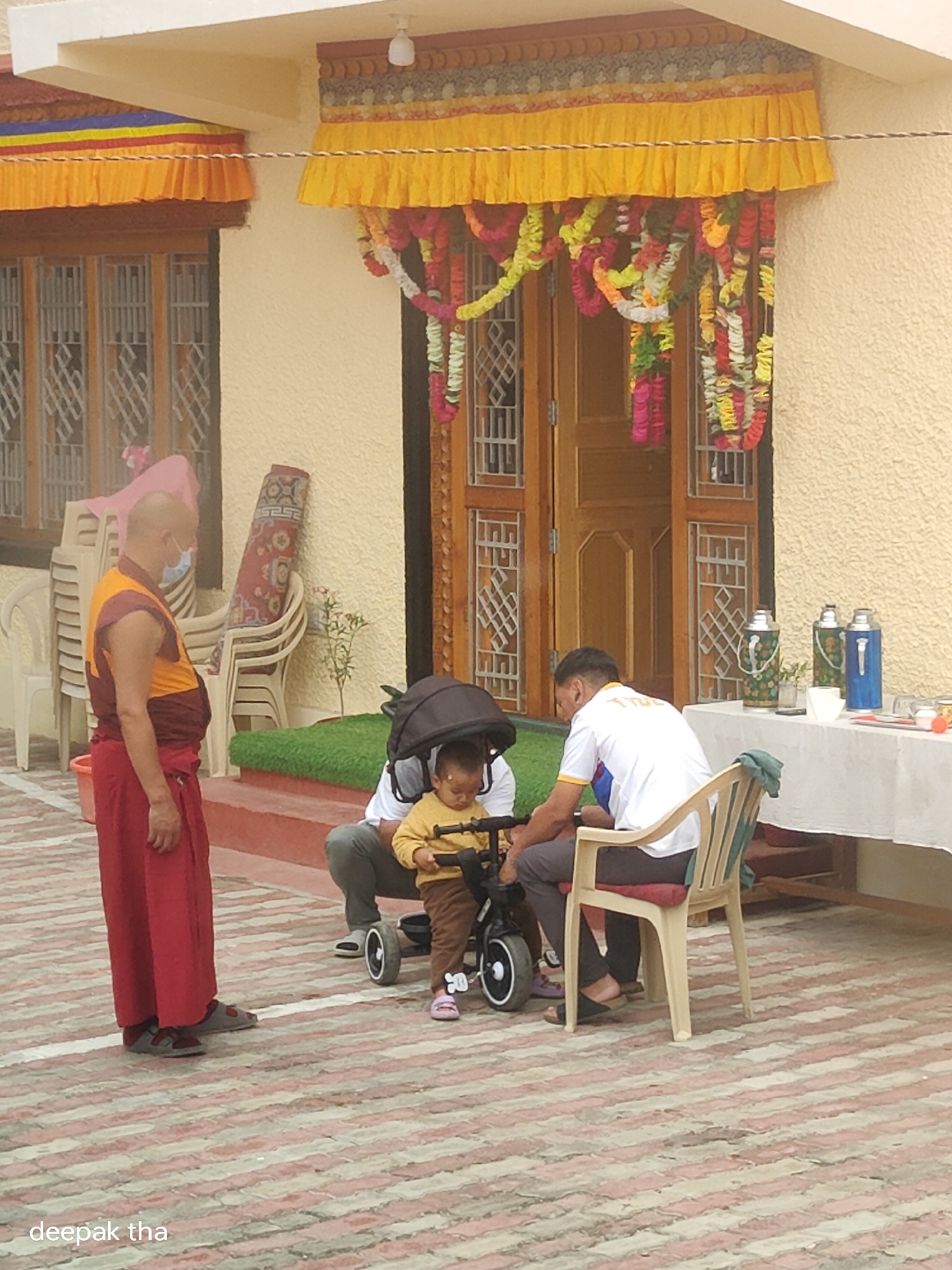 In Leh, India, in front of a monastery entrance adorned with yellow banners and colorful garlands, a Buddhist monk in traditional orange and maroon robes stands facing left. He wears a blue surgical mask. To his right, a man in a white t-shirt and dark pants crouches beside a toddler on a small black tricycle. The toddler, wearing a yellow shirt and brown pants, is holding the tricycle's handlebars. Behind them, a white tablecloth is spread over a table laden with three thermos bottles and some glasses. Stacked chairs are visible to the left, and a rolled-up rug leans against the building's textured yellow wall. The ground is paved with a pattern of pink and beige interlocking bricks.