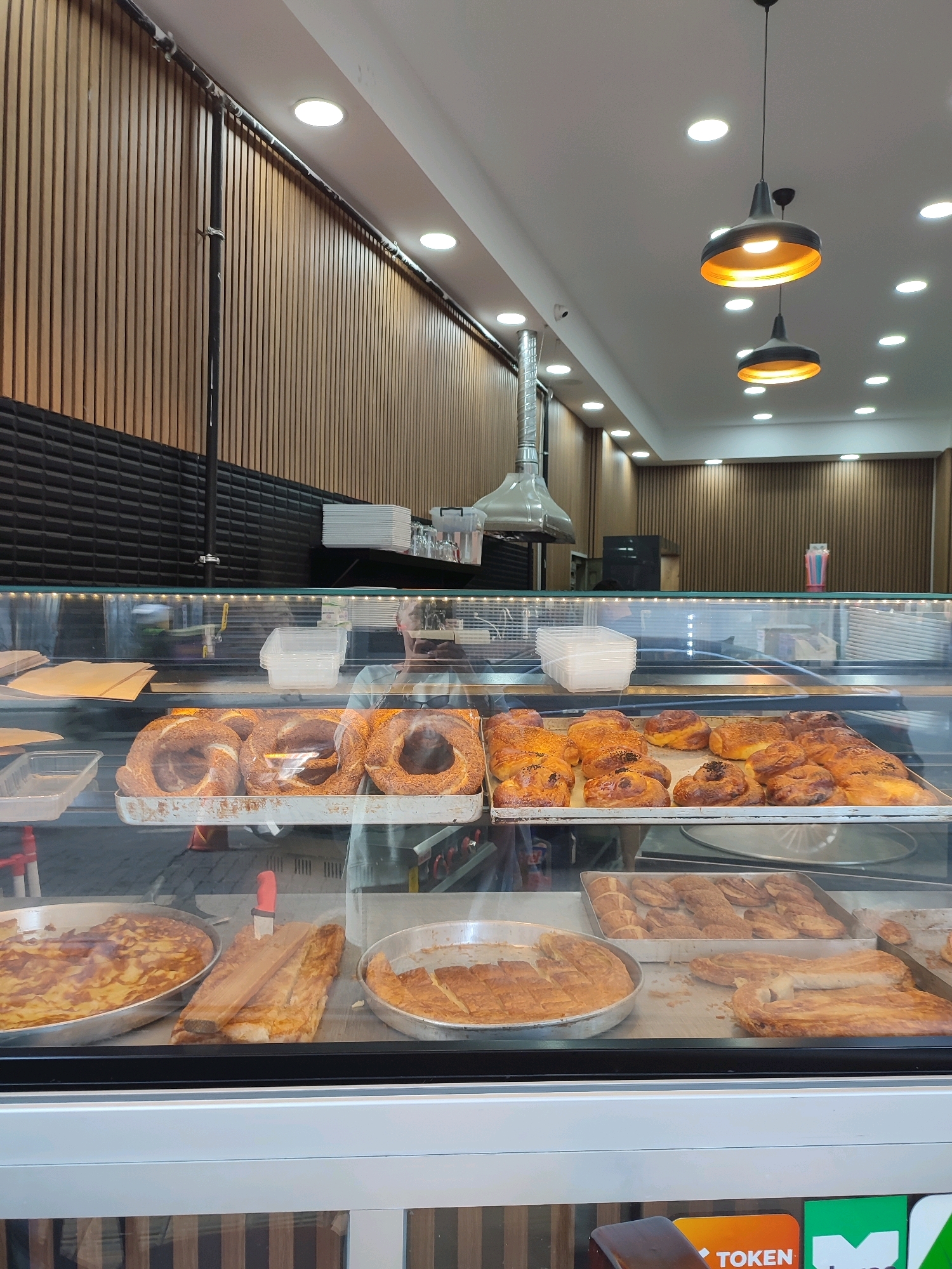 The image captures a display case filled with various baked goods, likely in a bakery. The display case is made of glass and is illuminated by overhead lighting. In the foreground, there are several trays of pastries, including circular, sesame-covered bread rings, and golden-brown, filled pastries. Behind these, more baked goods are visible, including what appears to be a large, flat pie or pastry. The background shows a wall with vertical wooden slats, a black textured panel, and some stacked white plates and glasses on a shelf. Two industrial-style pendant lights hang from the ceiling. The overall impression is of a well-stocked bakery counter. This scene is located in Kadıköy, Turkey.