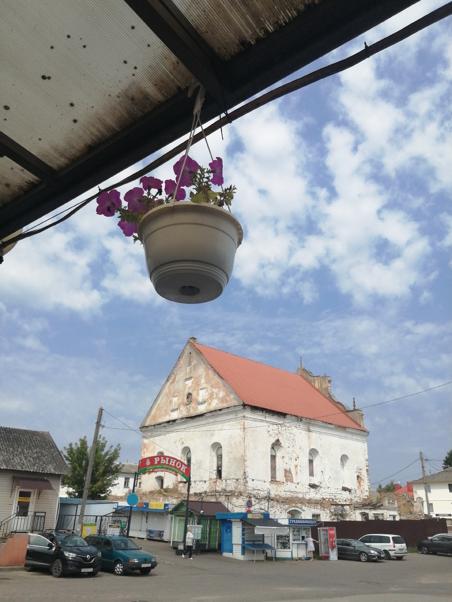 The image captures an outdoor scene in Slonim, Belarus, likely near a market given the signage and building structures. In the foreground, a white hanging planter filled with pink petunias is suspended from a corrugated metal overhang. Below, two cars are parked: a black Renault on the left and a dark green sedan on the right. Behind the cars, a building with a red roof and peeling white paint dominates the midground. A red and green banner with Cyrillic text reading "РЫНОК" (Market) is affixed to the front of this building, indicating its function. Several market stalls with blue and white accents are visible in front of the building. A woman in a white dress is standing at one of these stalls, interacting with a person inside. To the left of the main building, a smaller building with a grey roof is present. The background features a cloudy blue sky and more buildings in the distance, suggesting a developed urban or semi-urban environment. The overall atmosphere appears to be a typical day at a local market.
