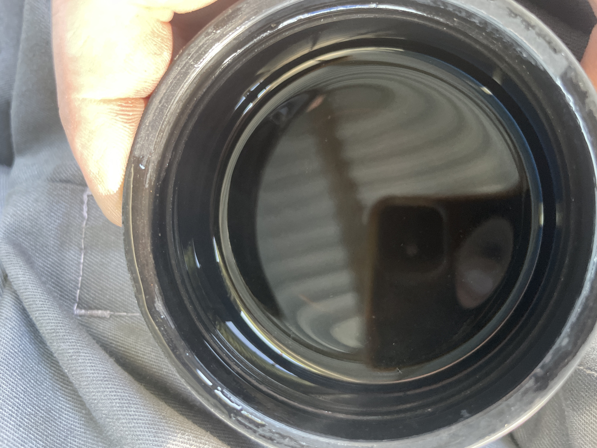 A close-up, overhead shot shows a black container filled with a dark, oily liquid. The container has ridged sides and a wide opening. The liquid appears viscous and reflects light, creating a swirling pattern on its surface. A person's hand, with visible fingerprints, rests on the rim of the container. The background consists of a textured, grey fabric, possibly clothing or upholstery. There are no visible signs of the city of Сокуры, Russia in the image.