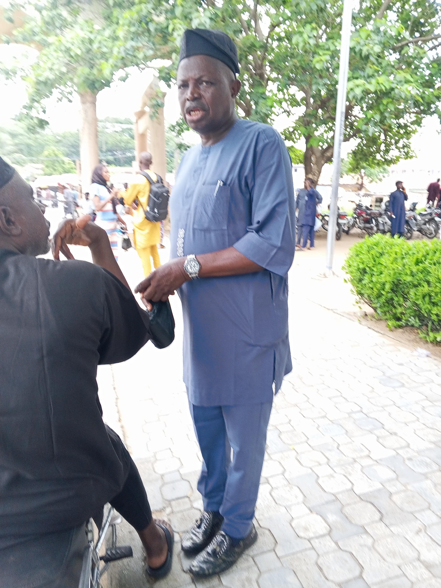 A man wearing a navy blue cap and a matching long-sleeved kaftan stands on a paved walkway. He has dark skin and is holding a black cloth in his right hand, near his waist. On his left wrist, he wears a silver watch. To his left, another person in a black outfit is visible from the back, seated in a wheelchair. In the background, several people are standing around, with motorcycles parked behind them. Trees with green foliage are present in the scene, and the ground is paved with interlocking bricks. The overall environment appears to be an outdoor urban setting in Lokoja, Nigeria.