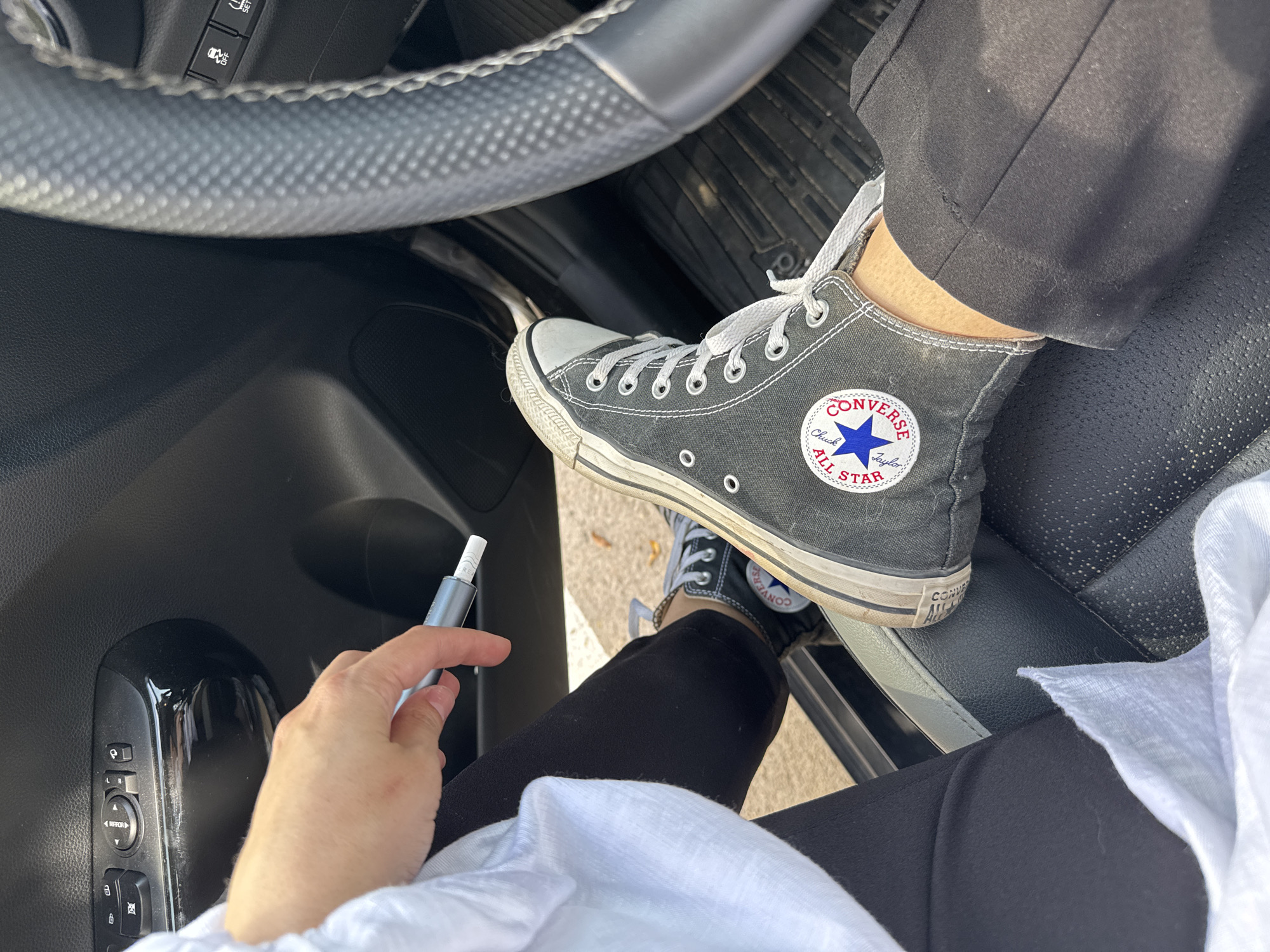 A person is seated in the driver's seat of a vehicle, holding an electronic cigarette device in their hand. The person is wearing black pants, a white shirt, and black Converse high-top sneakers. The vehicle's steering wheel and door controls are visible. The location appears to be indoors, possibly a parking garage, as indicated by the lack of strong direct sunlight and the interior of the vehicle. The environment is otherwise ambiguous, with no clear indicators of Tel-Aviv or Israel present in the image.
