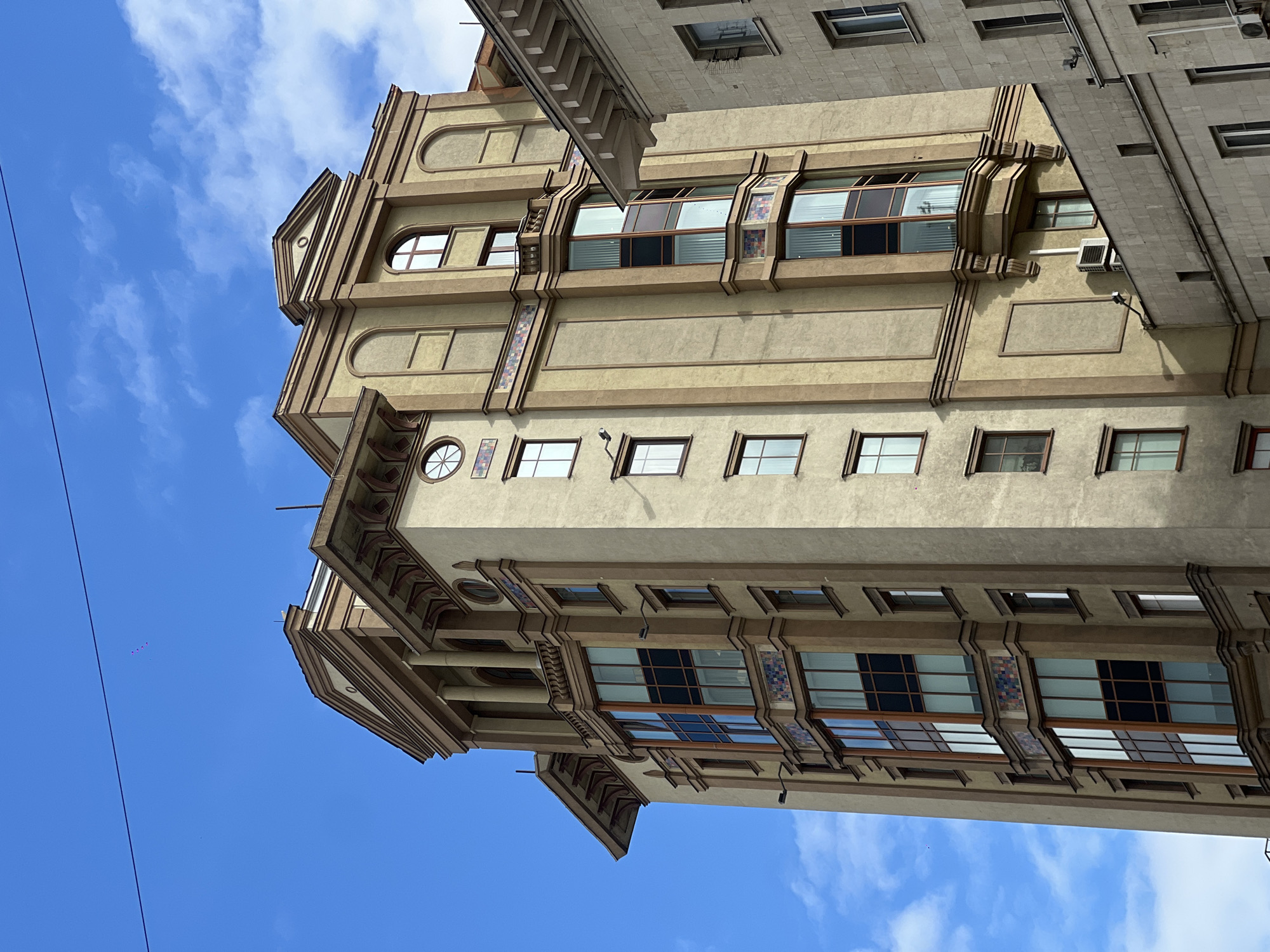 The image depicts a building in Moscow, Russia, specifically on Myasnitskaya Street. It is a multi-story structure with architectural details, including windows of varying shapes and sizes, decorative elements, and a light-colored facade. The sky is visible with a mix of blue and some clouds.