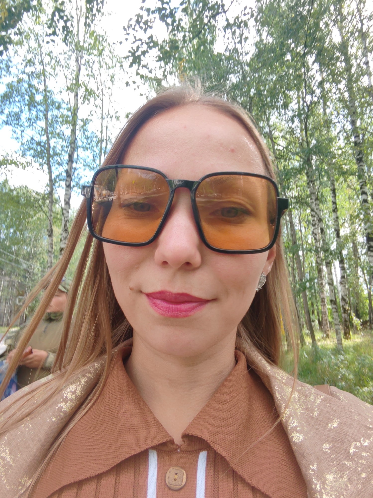 A young Caucasian woman with blonde hair is taking a selfie outdoors. She is wearing large, square-framed orange sunglasses, bright pink lipstick, and a brown collared shirt with gold detailing. Behind her, a forest of birch trees can be seen, with green foliage and dappled sunlight. A person in a camouflage jacket is visible in the background, to the left of the woman. The overall impression is a candid shot taken in a natural setting, likely on a sunny day. The presence of birch trees suggests a location in northern Russia, potentially in the city of Syktyvkar, which is known for its birch forests.