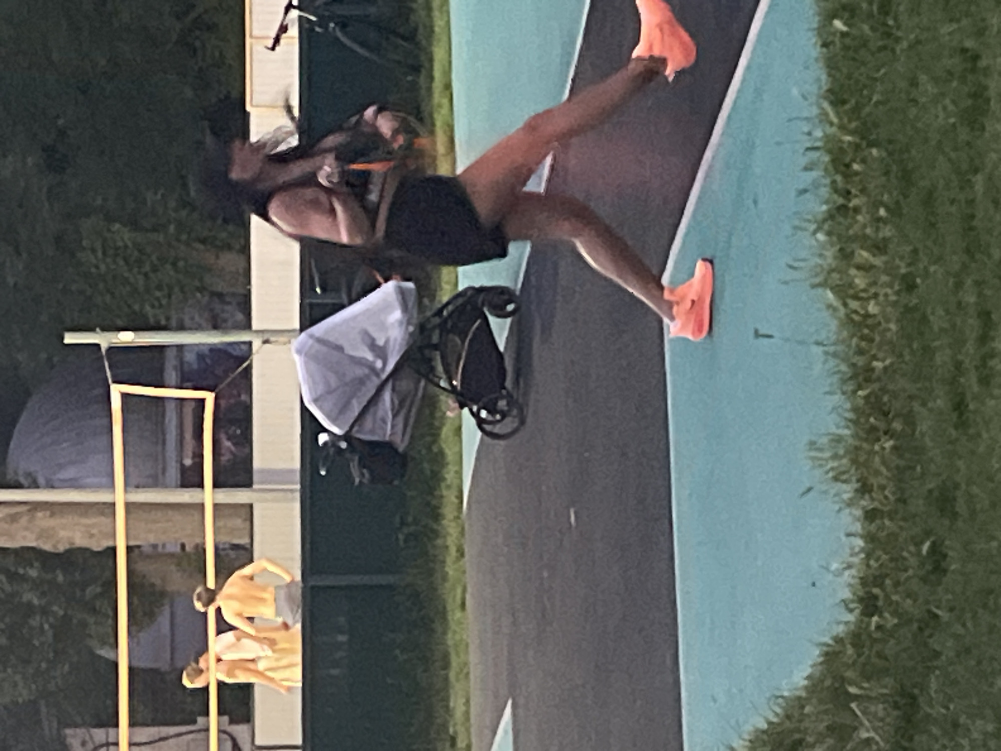 The image depicts an outdoor scene with a volleyball net in the background, suggesting a recreational area. In the foreground, a woman wearing a black baseball cap, a dark top, and black shorts is captured mid-stride, her orange athletic shoes prominent. She appears to be moving from left to right, with her right leg extended forward and her left leg trailing. A baby stroller with a grey cover is positioned behind her to the left. Further in the background, three shirtless individuals, presumably men, are visible on what appears to be a court, possibly playing volleyball. The ground is a dark surface with turquoise and white markings. The environment appears to be a park or sports complex, with grass and trees visible beyond the playing area. There is no indication of the specific location being in Krasnodar, Russia, as the image lacks any identifying landmarks or context to suggest this.
