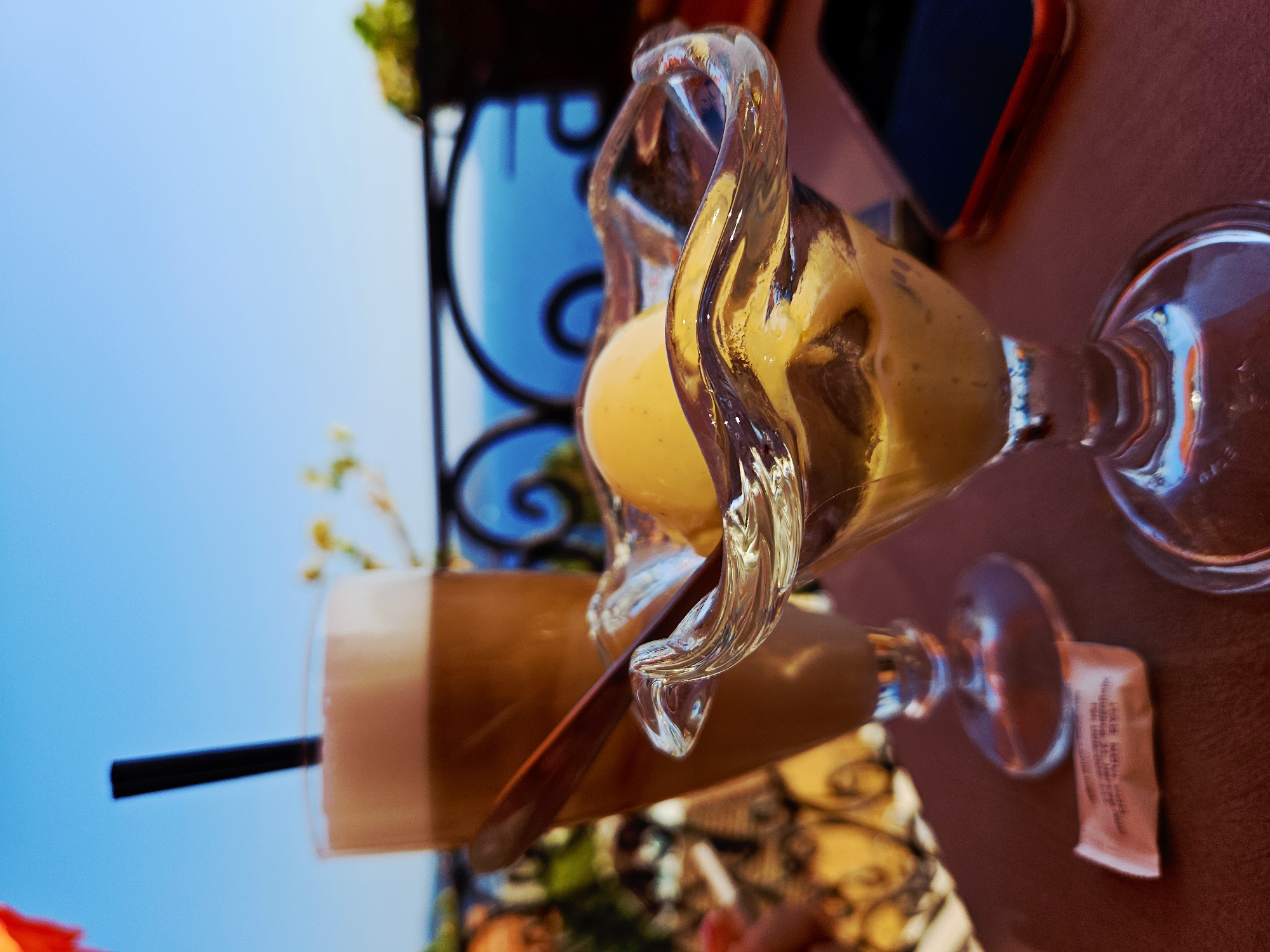 The image depicts a close-up view of a table at an outdoor cafe, with a backdrop of the Adriatic Sea and a clear blue sky. In the foreground, a glass of iced coffee with a black straw and a wooden spoon rests on a dark red surface. To the right of the coffee is a decorative glass dish containing a scoop of pale yellow ice cream, likely vanilla or lemon, with some creamy mixture underneath. A small packet, possibly sugar or sweetener, is on the table to the left of the coffee. A smartphone with a red case is partially visible to the right of the ice cream dish. The background reveals a wrought-iron railing overlooking a body of water, suggesting a seaside location, potentially a balcony or terrace in Herceg Novi, Montenegro, given the distinctive architectural style and the typical cafe setting in such tourist destinations. The lighting suggests it is daytime, likely afternoon, with sunlight casting shadows and highlighting the textures of the drinks and ice cream. No people are clearly visible, though there might be a hint of a hand or arm in the far left of the image, out of focus. The overall impression is one of leisure and relaxation, perhaps a moment of enjoying a sweet treat and a refreshing beverage with a scenic view.