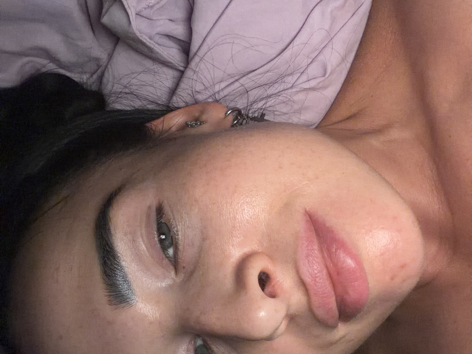 The media file displays a close-up, slightly rotated portrait of a person, likely female, from the neck up, lying down in an indoor setting, possibly a bedroom in Kokkorevo, Russia. The individual has dark hair, some strands of which are visible loosely spread on a light purple or lavender wrinkled pillowcase.
The subject's face is prominent, featuring an open eye with a neutral, calm gaze and a reflection in the pupil. Dark, well-defined eyebrows frame the eye. A nose and full, pinkish lips are also visible. The skin appears natural, with a dewy or slightly moisturized finish, and faint freckles are scattered across the cheek. Natural skin texture, including some minor pores and slight redness or blemishes, can be observed on the face and neck.
Notable details include multiple ear piercings on the visible ear. One piercing consists of a cluster of small, shiny studs or gems on the upper cartilage, while another, more prominent silver-colored earring, possibly a chain or linked hoops, is attached to the lower part of the ear. The overall expression conveys a sense of relaxation or contemplation. There is no visible text in the image. The time of day and external weather conditions are not discernible from this indoor close-up.
