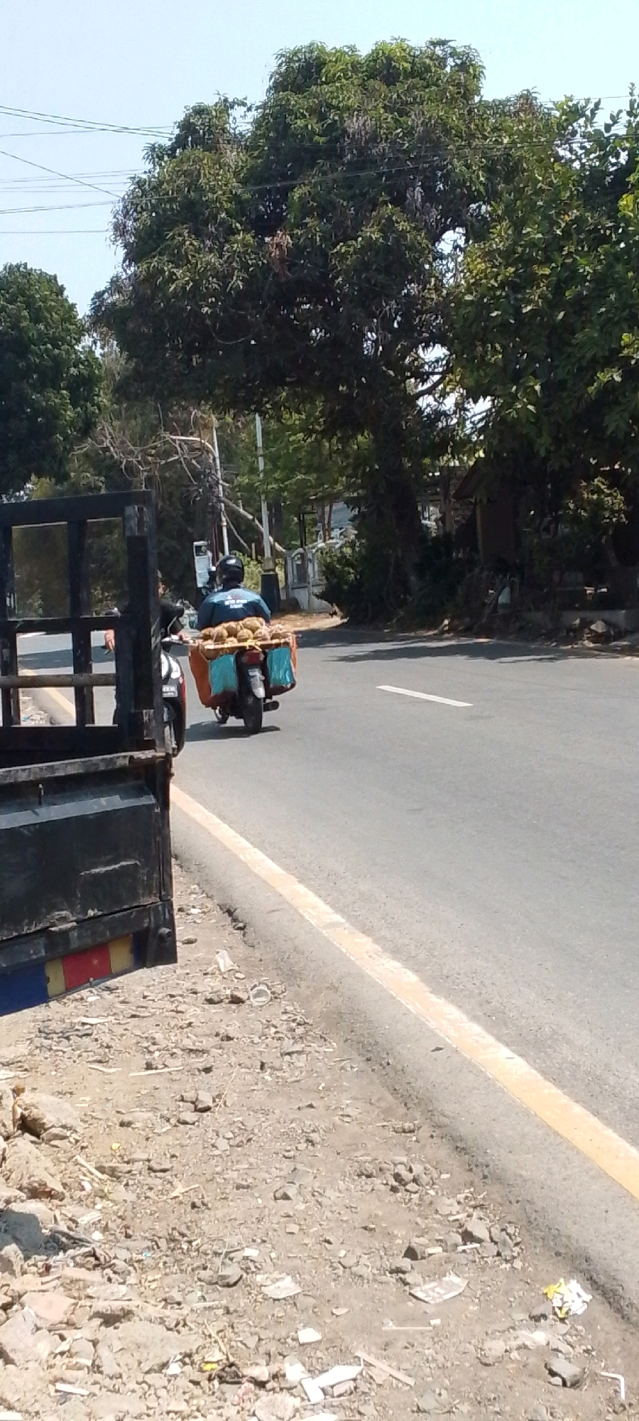 In Cilaku, Indonesia, a person wearing a blue shirt and a black helmet rides a motorbike down a paved road. The motorbike is loaded with durians, a fruit, in the back. The motorbike is being followed by a black truck. The scene is set against a backdrop of trees and a clear sky. The road has a yellow dividing line and is bordered by a dirt shoulder littered with debris.