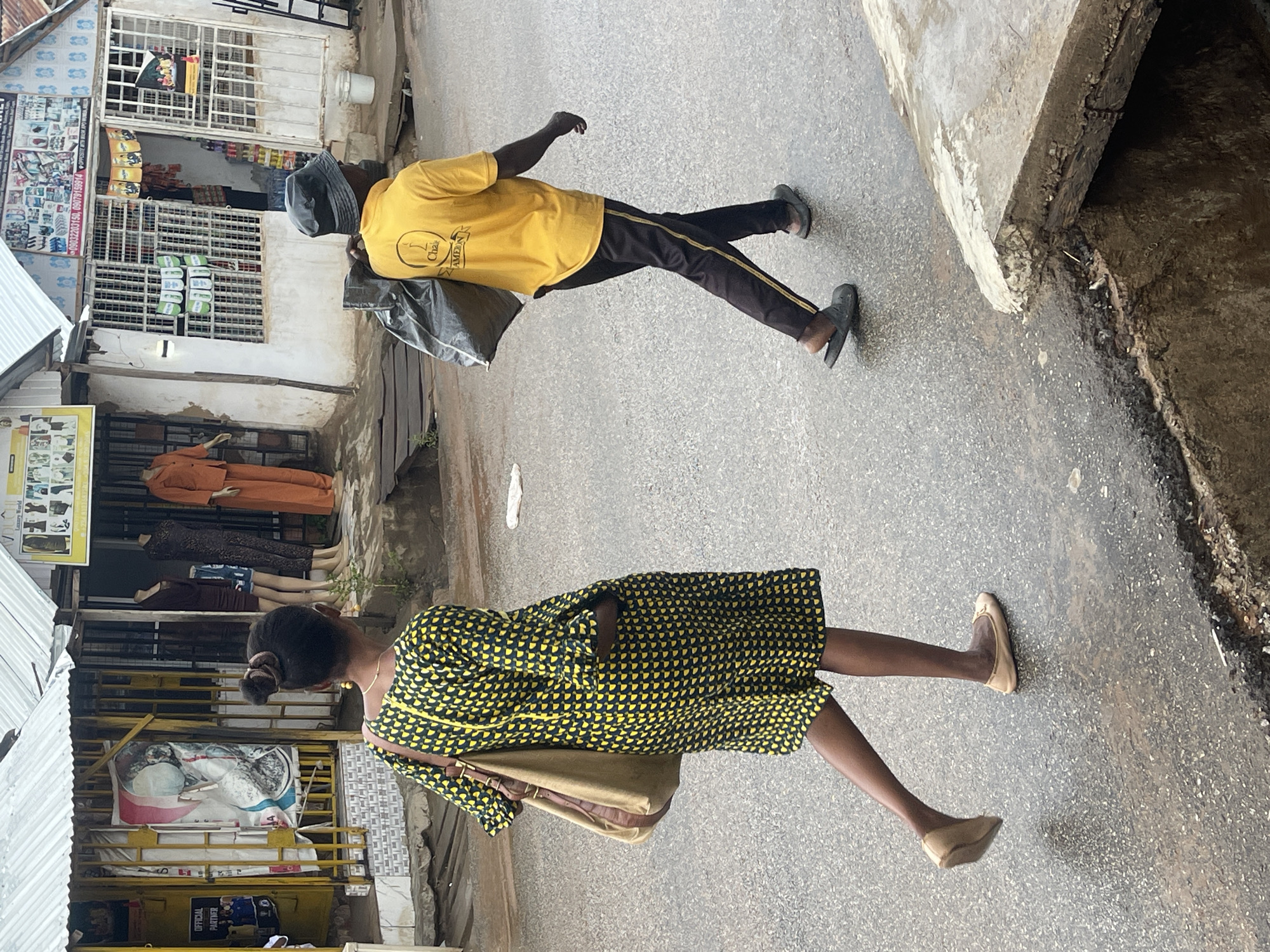 This media file captures a vibrant outdoor street scene in Abuja, Nigeria, under bright daylight. Two individuals are depicted walking on a paved road, moving away from the viewer.
In the foreground, a person wears a black and yellow patterned dress, light-colored flat shoes, and carries a light brown shoulder bag. Their hair is styled in a bun, and a necklace is visible. Further along the street, another person wears a yellow t-shirt with a logo and text on the back that reads "CH2 AMERICA," dark trousers with a yellow stripe, dark flat shoes, and a dark cap. This individual carries a black bag slung over their shoulder.
The street is flanked by various commercial structures. On the left side of the scene, a shop with a white metal gridded security gate showcases products like bottles and packets inside. A sign on its wall displays images of electronics, alongside text including "SHOP" and phone numbers such as "09091599814." Below this, another shop area features mannequins dressed in different outfits, including an orange jumpsuit and a dark patterned dress. Nearby, a sign reading "OFFICIAL PARTNER" with what appears to be a football club logo is visible. The ground is a mix of asphalt or concrete road, with a rougher, darker section along one edge, possibly a drain or unpaved area. The scene depicts typical daily street activity in an urban or semi-urban Nigerian setting during clear, sunny weather.