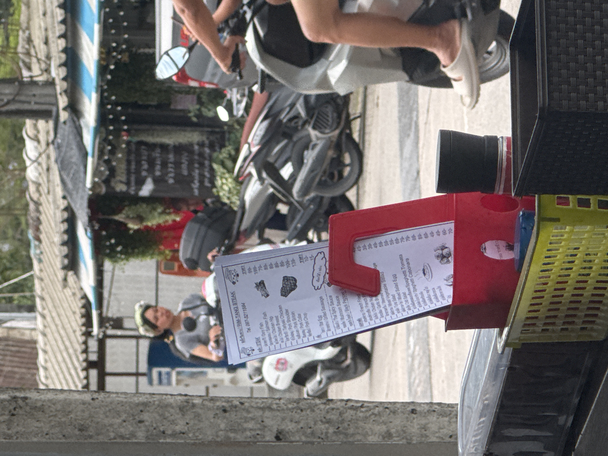 This is a medium-shot, eye-level, outdoor photograph taken in Kathu, Thailand. The foreground is dominated by a red plastic holder containing a menu and a black plastic basket. Behind these, a person on a scooter is partially visible, with their legs and feet in white sandals evident. The scooter is silver and has a black seat. Further in the background, other scooters are parked, with one on the left being black and another more centrally located being silver with a red seat. A woman wearing a headscarf is seated on the black scooter. A building with a corrugated metal roof and blue and white striped awnings is visible in the mid-ground, and a concrete pillar is in the left foreground. The overall scene depicts a street-level view with a focus on the scooter and food stall elements.