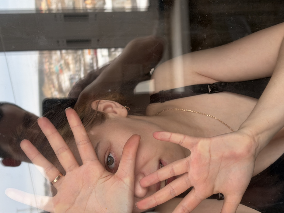 The image depicts an indoor scene in Chaikovsky, Russia, looking through a reflective glass window during daylight hours in winter. A fair-skinned adult female, positioned indoors, has both hands pressed against the window pane, partially obscuring her face. Her left hand, adorned with a gold-colored ring on the fourth finger, covers her upper face, leaving one green/hazel eye visible. Her right hand covers her lower face, revealing her mouth with visible teeth and what appear to be braces. She wears a black bra or tank top, a thin gold-colored chain necklace, and a small gold-colored hoop earring in her right ear. Reflected in the glass behind her is the indistinct upper head and shoulder of an individual, likely male, holding a dark smartphone, suggesting they are capturing the image. Beyond the window, a blurry, snow-covered urban landscape with buildings of varied roof colors and distant hills is visible under a bright sky.