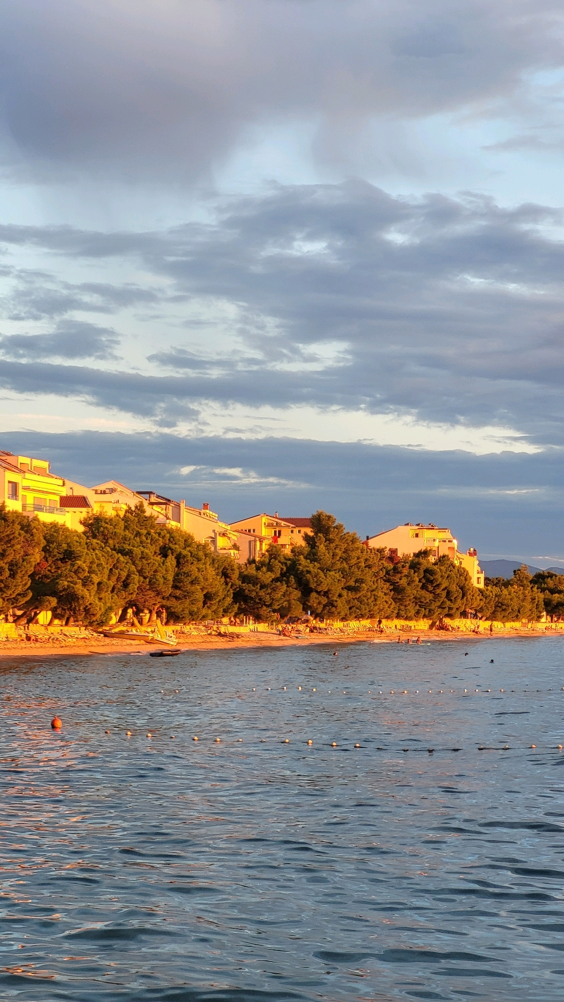 The image captures a serene beach scene at sunset in Općina Tučepi, Croatia. The golden light of the setting sun bathes the buildings and trees lining the shore in a warm glow. The sky is adorned with dramatic, textured clouds, a mix of light grays and blues.
In the foreground, the calm sea reflects the sky and the warm light. Small, rounded buoys are strung together, marking a boundary in the water. A single orange buoy floats independently. A few people are visible in the water, some swimming, one on a paddleboard, and another seemingly wading near the shore.
The middle ground showcases a sandy beach dotted with lounge chairs and umbrellas, suggesting a popular tourist destination. A small group of people are present on the beach, some sitting, others possibly strolling. Lush green trees form a dense line between the beach and the buildings, providing a natural barrier.
Behind the trees, a row of modern, multi-story buildings with light-colored facades stretches along the coastline. Their windows and balconies are visible, some reflecting the sunset. The architecture appears to be contemporary residential or hotel structures. The overall impression is one of a peaceful and picturesque coastal resort town during the late afternoon or early evening.