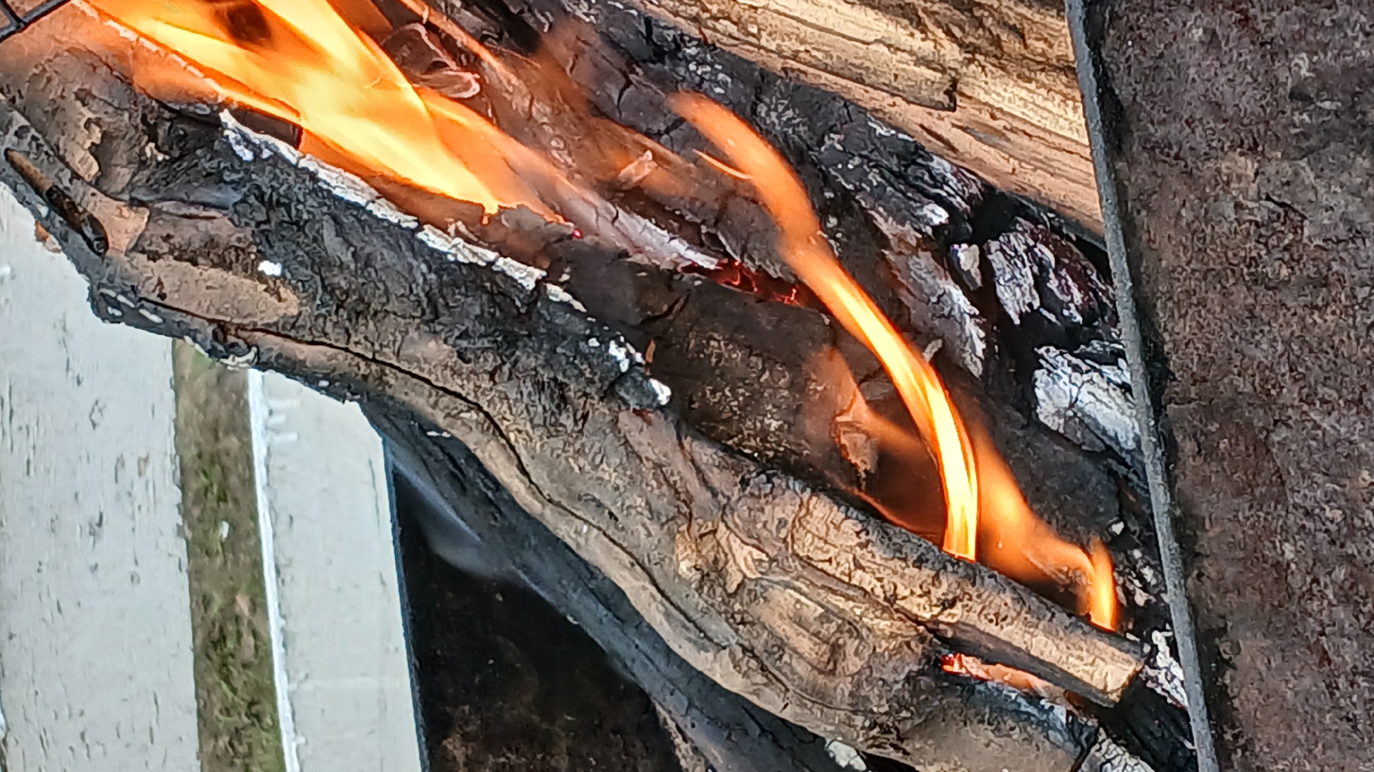 A close-up, high-angle shot shows logs burning in a metal brazier. The logs are dark gray and charred, with visible cracks and rough textures. Orange flames lick around the wood, creating a dynamic and warm glow. The brazier is made of dark, rough metal, and appears to be filled with ash and embers at the bottom. In the background, a white structure with peeling paint and a strip of green grass are visible, suggesting an outdoor urban setting. The image was taken in Derköl, Kazakhstan.