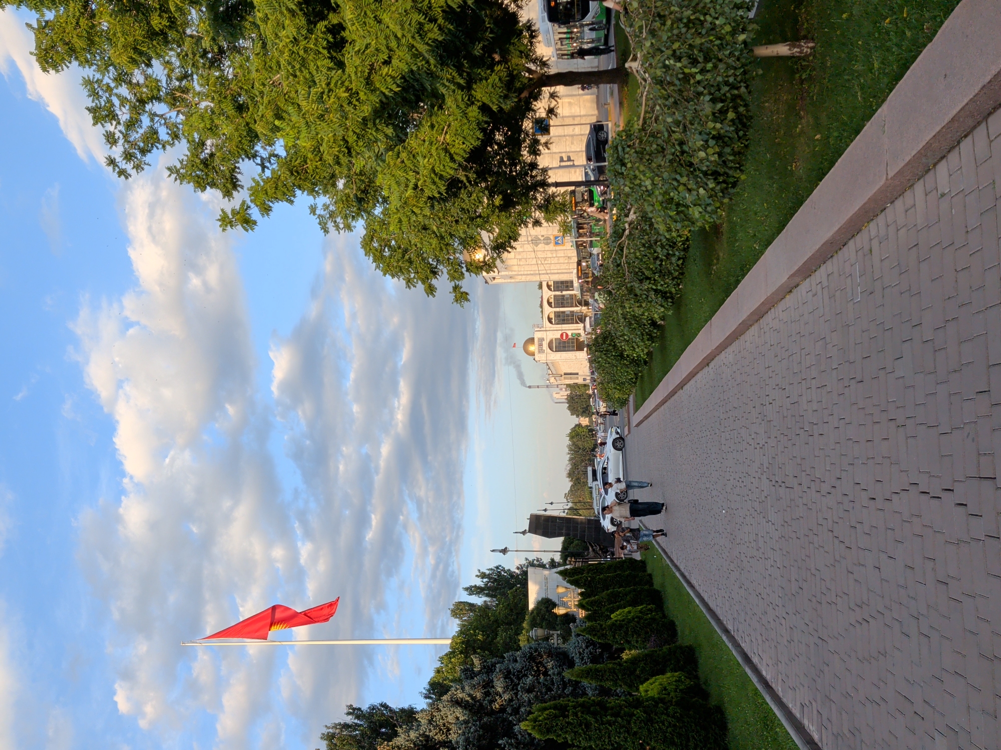 A vibrant red flag flutters on a white flagpole against a backdrop of a partly cloudy blue sky. To the right, a tree with lush green foliage is in the foreground, casting shadows. Beyond the tree, a paved walkway with a textured surface stretches into the distance, bordered by manicured green grass. In the middle ground, several people are gathered near a white car parked on the side of a road. The road appears to be a busy thoroughfare with vehicles, including buses, visible. In the background, modern buildings with architectural details stand under the soft light of what appears to be late afternoon or early evening. The overall scene suggests an urban setting, possibly a public square or park in Bishkek, Kyrgyzstan, on a pleasant day. The mood is calm and observational, capturing a slice of everyday life in the city. There is no visible text in the image.