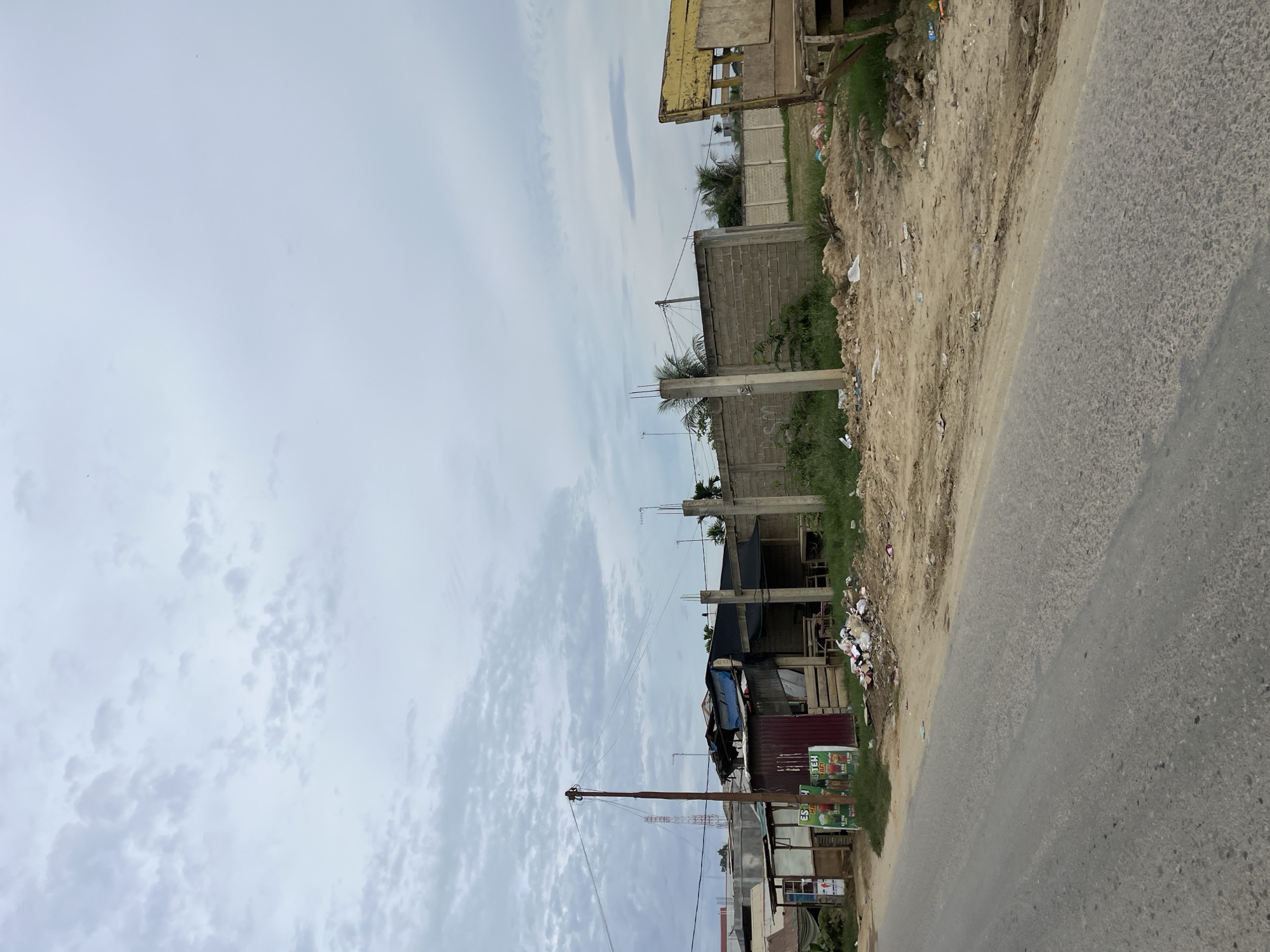 A roadside scene in Landuh, Indonesia, shows a mixture of structures and debris under an overcast sky. A paved road occupies the foreground, with a dirt shoulder bordering it. The shoulder is strewn with trash, including plastic bottles and paper.
Several small, makeshift buildings are visible on the left side of the road. One appears to be a small shop with colorful posters advertising drinks. Adjacent to it are structures with corrugated metal walls and tarpaulin roofs, some in disrepair. A wooden pole with electrical wires stands prominently near these buildings.
Further into the scene, a low, grey brick wall runs horizontally. In front of the wall, several concrete pillars stand at intervals, some with rebar protruding from the top, suggesting ongoing or incomplete construction. Behind the wall, more buildings and greenery are visible, including palm trees. On the far right, part of a weathered yellow truck or trailer is visible, its wooden structure exposed. The ground is uneven, with patches of dry grass and exposed soil.