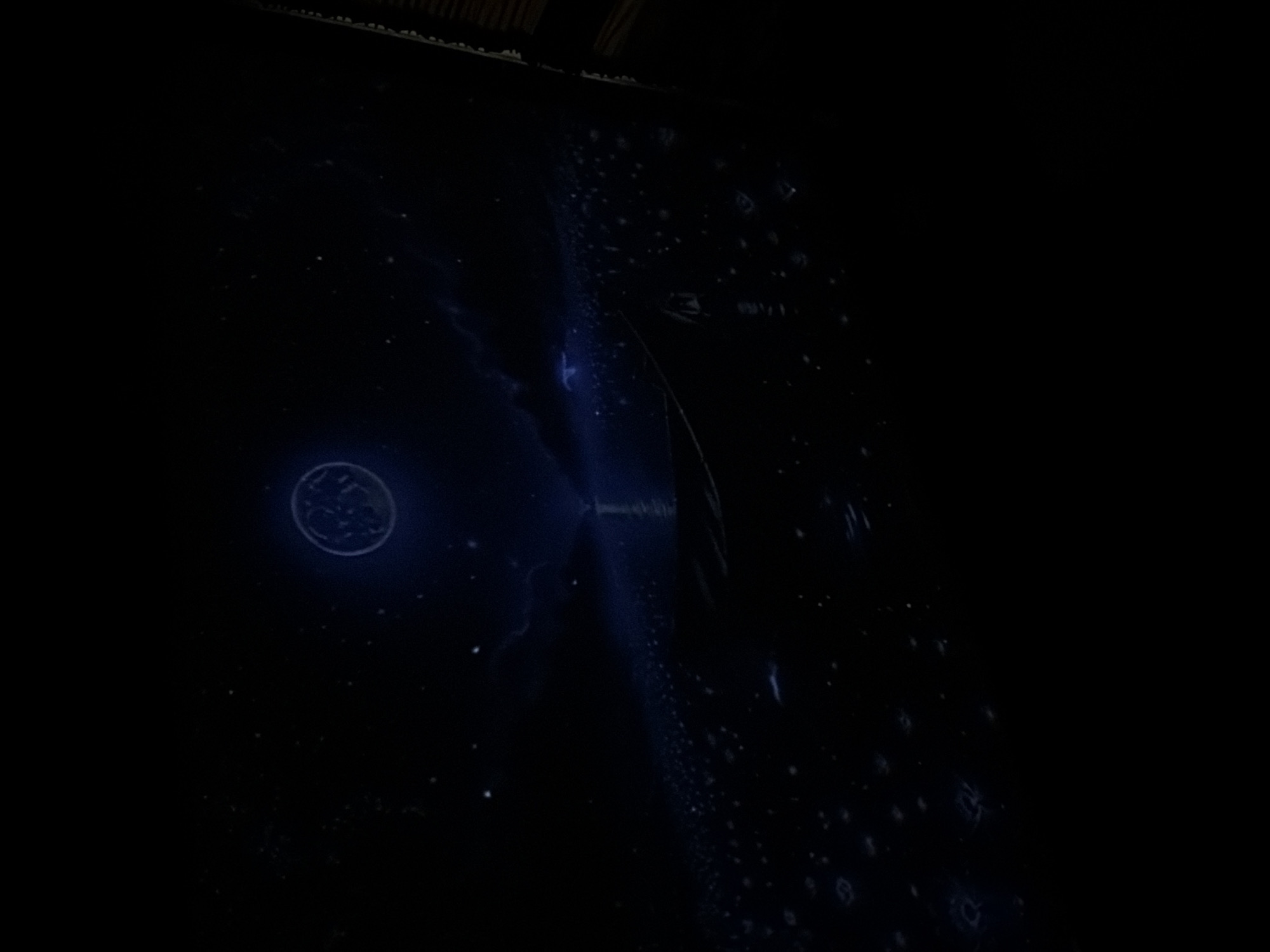 The image depicts a dark, atmospheric scene that appears to be a digitally rendered or artistic representation rather than a photograph of a real location. A large, luminous, disc-shaped object, possibly a moon or a celestial body with intricate patterns, hangs in a star-speckled night sky. Below the sky, a vast, dark body of water is visible, reflecting faint blue lights from the sky. In the foreground, a small boat with a visible silhouette is positioned on the water, with its bow pointing towards a distant light source that creates a beam extending across the water. The water's surface is dotted with numerous small, glowing blue shapes that resemble abstract symbols or glyphs. The right edge of the image shows a portion of a textured, wooden structure, suggesting it might be a framing element or part of a larger composition. There is no indication of Tbilisi, Georgia, or any identifiable urban elements within this image.