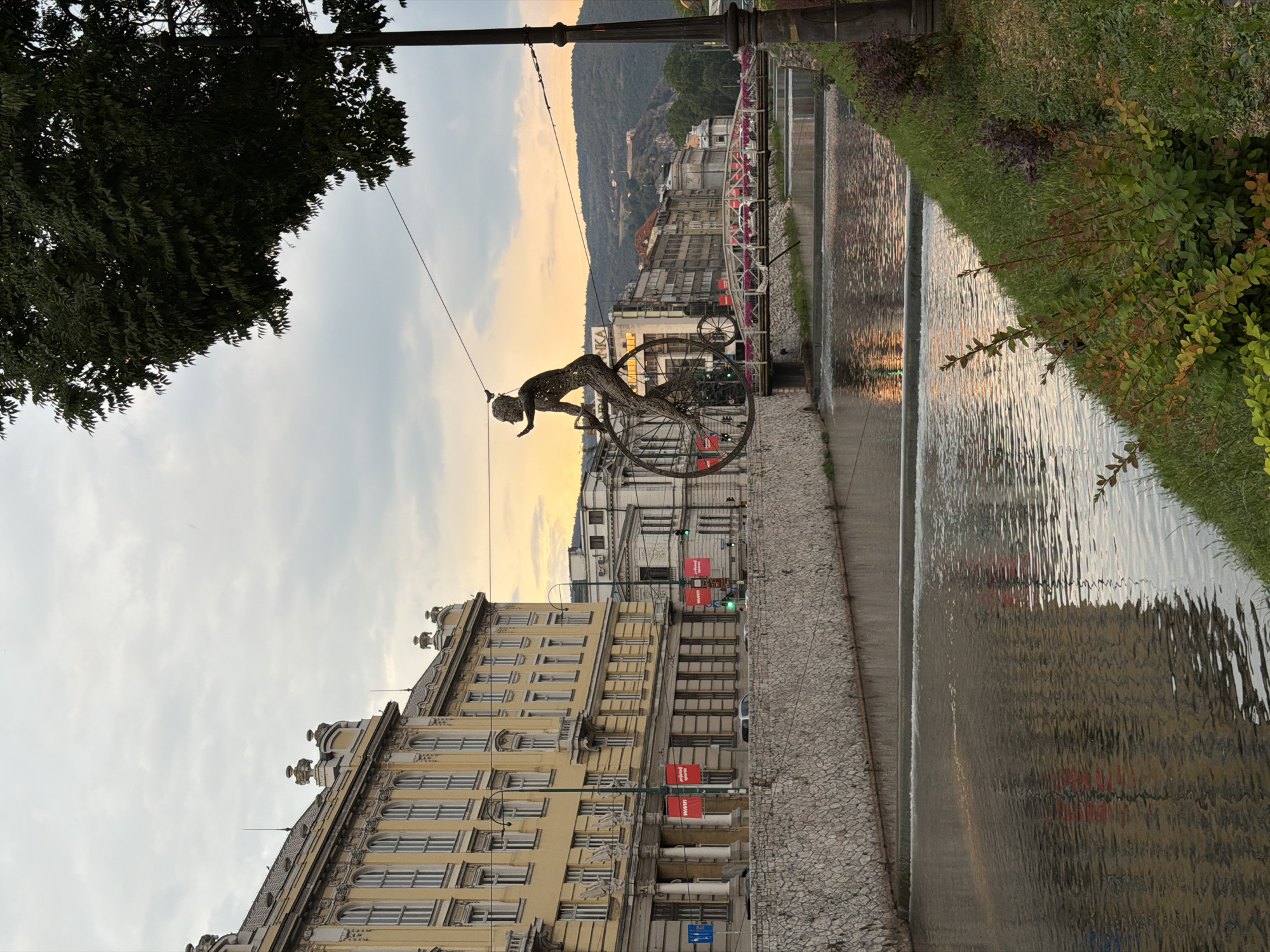 A sculpture of a cyclist in motion is the central subject, suspended in mid-air above a street. The cyclist is crafted from metal, with intricate detailing suggesting movement and effort. Behind the sculpture, buildings line the street, with a mix of architectural styles visible, some appearing older and ornate, others more modern. A canal or river runs alongside the street, its surface rippling and reflecting the warm tones of the sky. Greenery, including trees and bushes, frames the scene, adding a touch of nature to the urban environment.
The time of day appears to be either sunrise or sunset, given the soft, golden light illuminating the sky and reflecting in the water. The sky itself is a blend of soft blues and oranges, with clouds present. The weather seems calm and pleasant.
In the background, a hilly landscape is visible, dotted with more buildings, suggesting the location is nestled within a valley or at the edge of a city. The scene is tranquil, with a sense of stillness, broken only by the implied movement of the cyclist sculpture.
There are several pieces of visible text on red banners attached to lampposts along the street. Though small and somewhat blurry, they appear to contain the word "MARKET" or a similar word in English, repeated. A green traffic light is also visible in the distance. The overall impression is one of a peaceful urban setting during the twilight hours, enhanced by the artistic presence of the cyclist sculpture and the natural beauty of the water and sky.