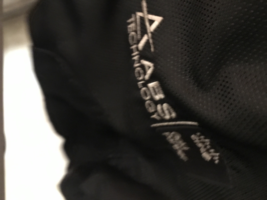 The image displays a severely out-of-focus, close-up view of black fabric with a white label or embroidery. The fabric exhibits a fine, perforated or mesh texture in areas. The legible text on the label, oriented vertically and largely upside down, includes "AXLABS TECHNOLOGY" and "ABS". Additional smaller, white text is visible but illegible. The upper portion of the frame shows a blurred white area, likely an overhead light source or a bright surface. There are no individuals, discernible actions, or environmental details present to determine a specific location within Calabar, Nigeria. The scene is static, focused solely on the textile detail.