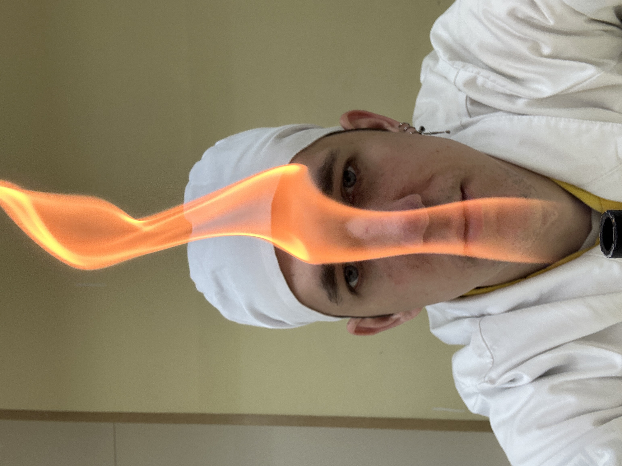 A person wearing a white surgical cap and jacket, possibly a medical professional, is holding a lit blowtorch. The person's face is partially obscured by the flame. The location is not identifiable.