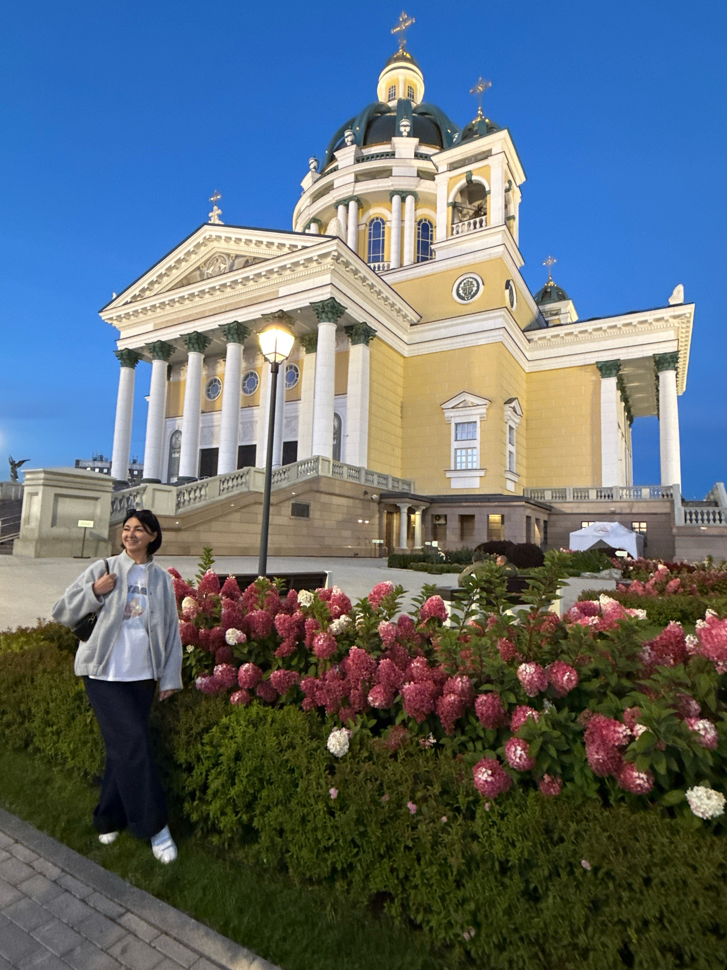 A woman stands in front of a large, yellow, neoclassical-style church with a domed roof. She is wearing a grey cardigan over a white t-shirt and dark pants, with white shoes. She is holding a black bag on her left shoulder and smiling. In front of her is a garden bed filled with pink and white flowers. The church is lit up and the sky is a deep blue, suggesting it is dusk or evening. This building is the Holy Trinity Cathedral located in Chelyabinsk, Russia.