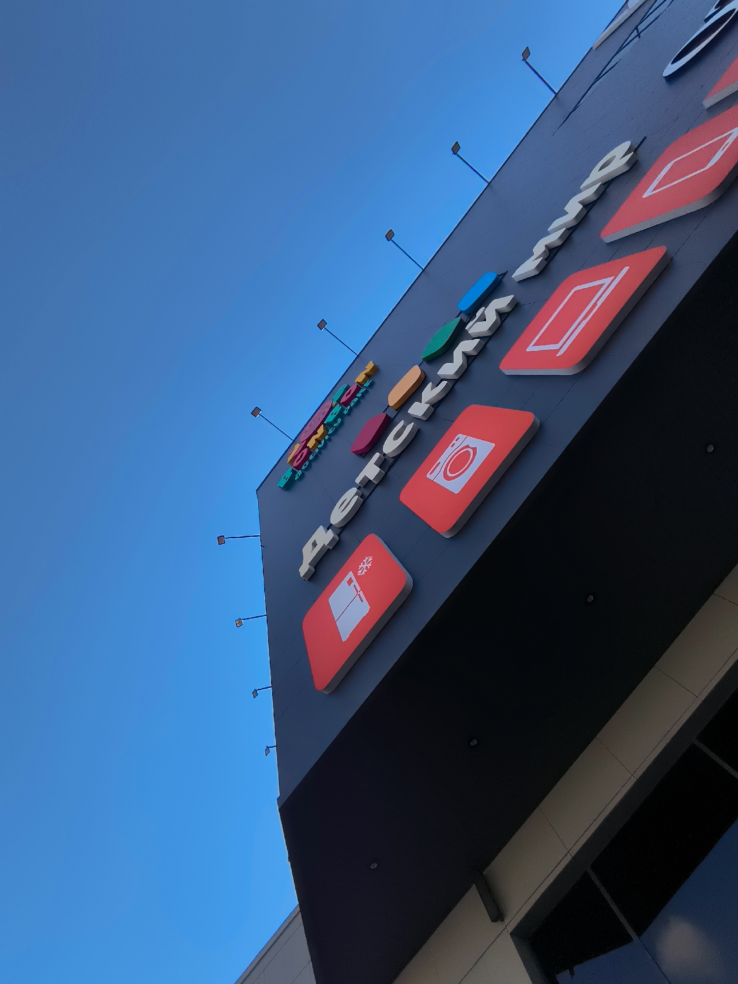 The image is a low-angle shot of the exterior of a modern building, likely a shopping center or commercial establishment, against a clear blue sky. The building's facade is dark gray and features prominent, three-dimensional signage. The signage consists of white, block-style letters spelling out "ДЕТСКИЙ МИР," which translates to "Children's World" in Russian. Interspersed with the letters are several brightly colored, rectangular icons on red backgrounds, each depicting a simple graphic. These icons include what appears to be a snowflake, a washing machine or similar appliance with a circular opening, and a rectangular outline, possibly representing a screen or a shelf. The building also has several protruding elements, possibly lights or decorative fixtures, on thin poles extending from the facade. The lower part of the image shows a glimpse of the building's structure, with bands of light-colored siding and dark windows. The perspective suggests the photograph was taken from ground level, looking upwards at the signage and the sky. The overall impression is one of a retail establishment designed to attract families, with a focus on children's goods. The location information provided indicates this building is situated in the city of Qarağandı, Kazakhstan.