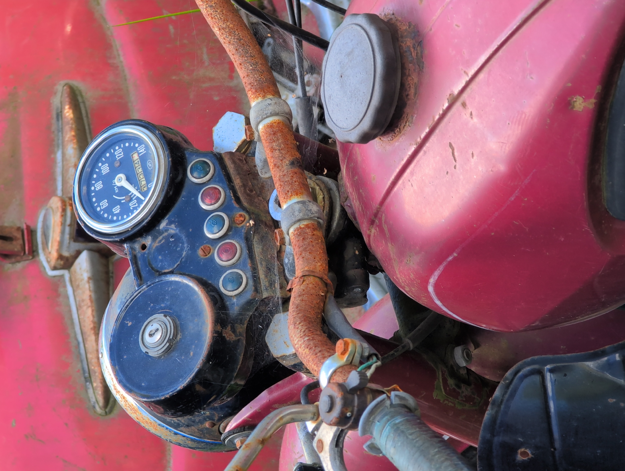 The image displays a close-up, angled shot of the front portion of a vintage, red motorcycle, likely located in Novoe Devyatkino, Russia. The motorcycle is in a state of disrepair, with significant rust evident on various metal components, including the handlebars and parts of the frame. The focus is on the dashboard, which features a circular speedometer with white markings and a needle pointing towards 20 KM/H. An odometer displays "37334" kilometers. Several circular buttons, some red and some blue, are visible below the speedometer. The motorcycle's red fuel tank is also partially visible in the foreground, showing signs of wear and tear. The overall environment suggests an outdoor setting, possibly a neglected area or a yard, with the background being a blurred expanse of red, consistent with the motorcycle's body. There are no people visible in the image.