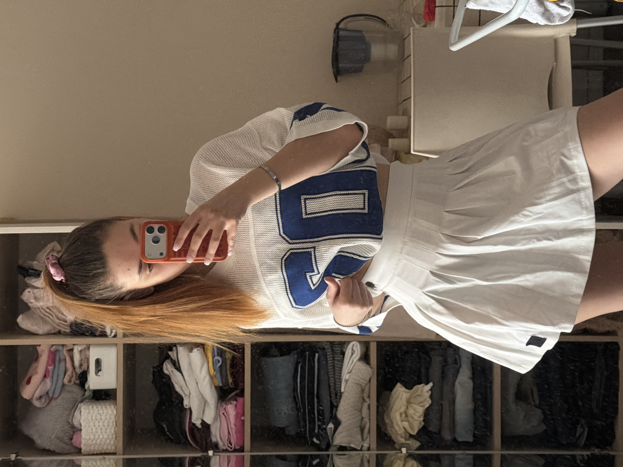 A young person with long, reddish-brown hair partially pulled back with a pink scrunchie is taking a mirror selfie. They are wearing a white mesh jersey with blue accents and a matching white pleated skirt. The person is holding an orange phone in their right hand, obscuring part of their face. The background reveals a closet system with open shelves containing assorted clothing items. Behind the person, there is a cabinet with items on top. The location appears to be indoors, likely a bedroom or dressing area. There is no visible indication of the city of Sofia, Bulgaria.