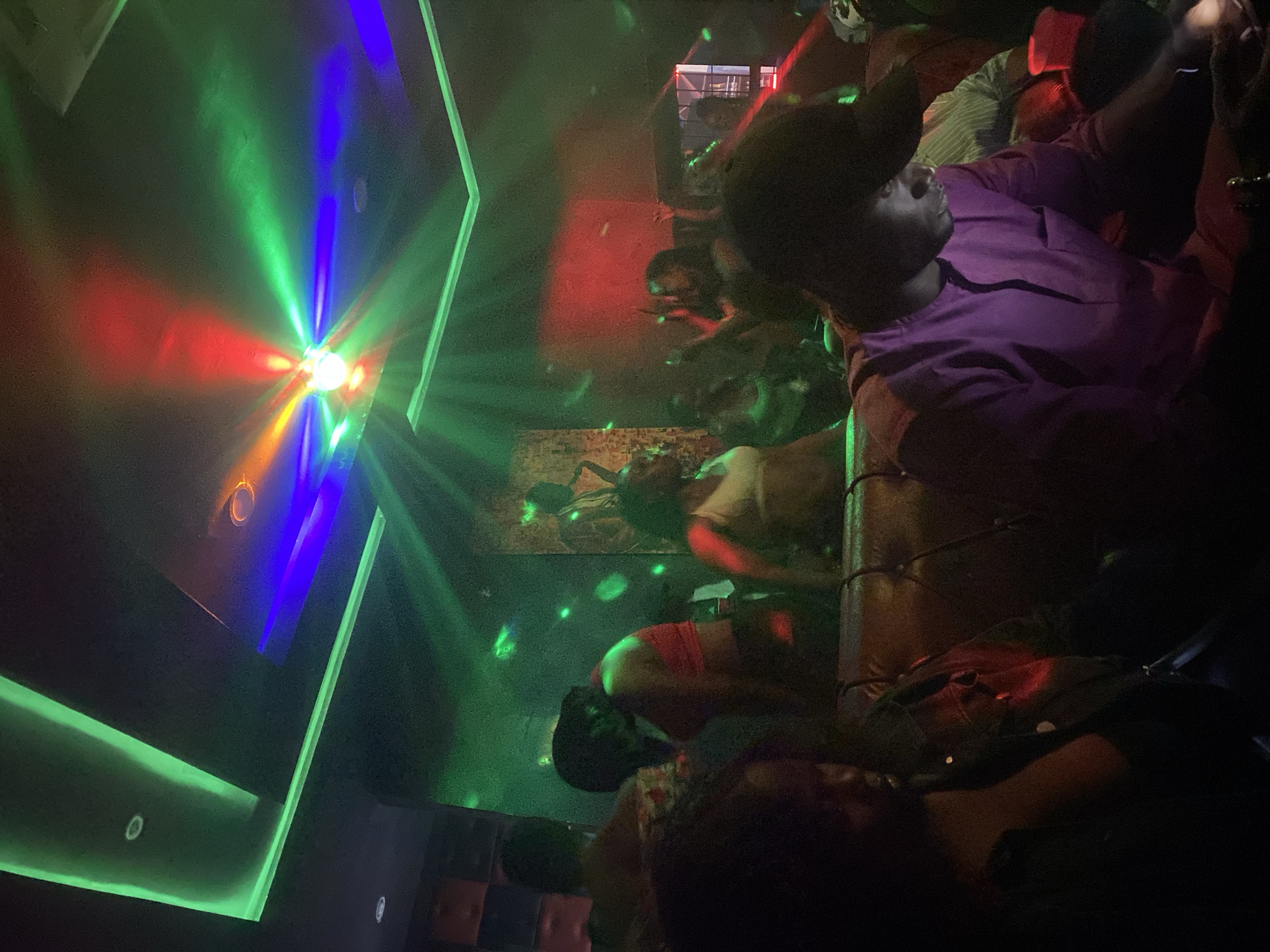 The image depicts an interior scene, likely a bar or lounge, in Gombe, Nigeria. Several individuals are present, some seated on a sofa, and others standing or moving around. A man in a purple shirt and a cap is prominently visible. The environment is dimly lit with various colored lights, including green, blue, and red, creating a vibrant, possibly party-like atmosphere. A painting or decorative piece is on the wall. The ceiling has green neon light strips.