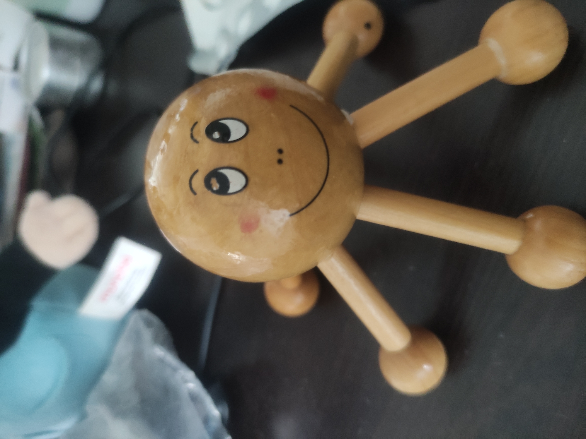 A wooden octopus-shaped massage tool with a painted face is placed on a dark, flat surface. The tool has a round head with a smiling face, rosy cheeks, and two black dots for a nose. It has six wooden legs with rounded ends. In the background, out of focus, a blue object, possibly a plush toy, and a white tag are visible. The environment appears to be indoors, on a desk or table. There is no indication of the location being in Baku, Azerbaijan.