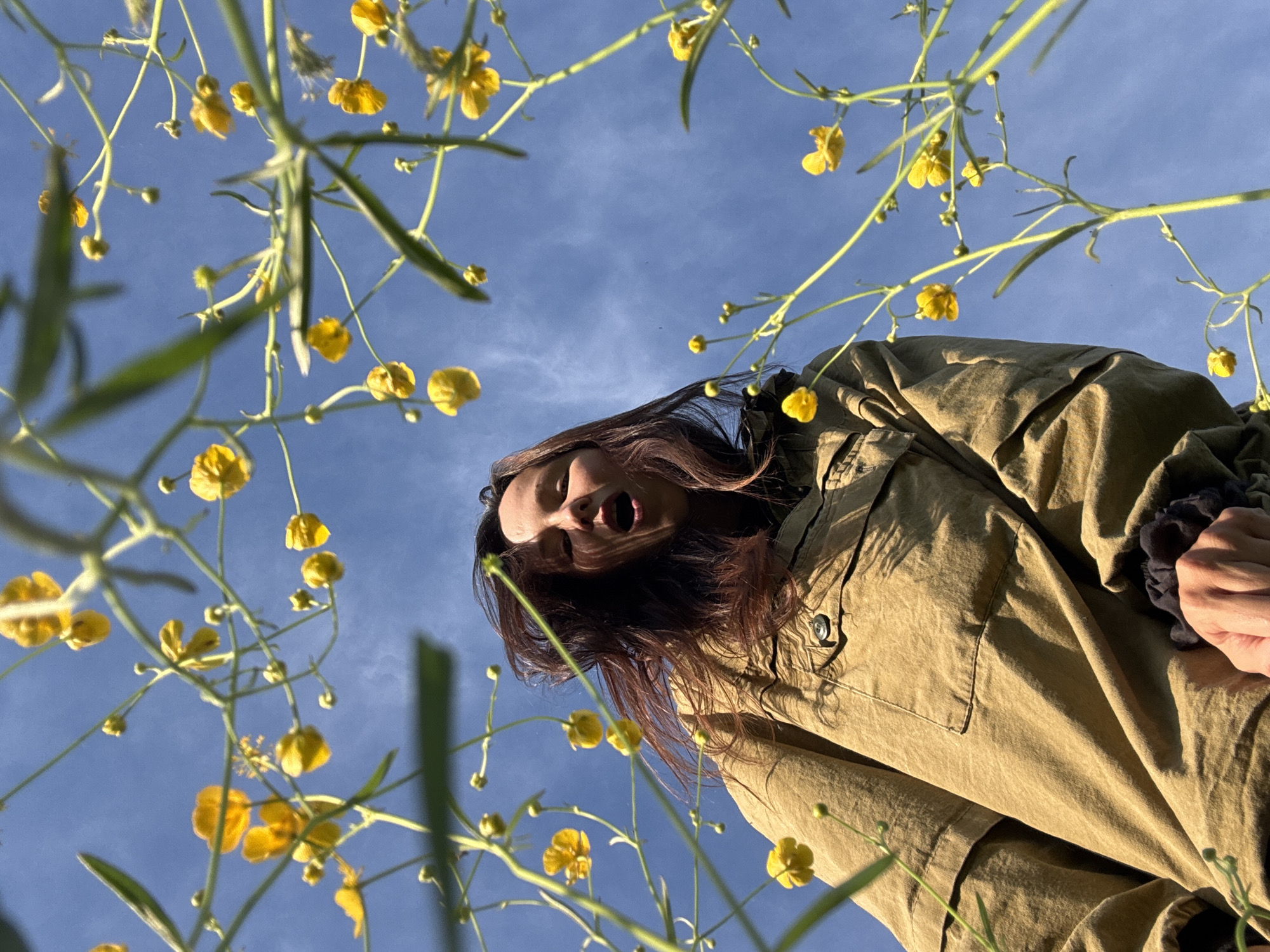 A low-angle shot captures a person in an olive-green jacket looking down at the camera, with their mouth open. The background is a blue sky dotted with small, yellow, buttercup-like flowers on delicate stems. The setting seems to be a field of these flowers. There's no explicit information indicating that this scene takes place in the city of Egorii, Russia. The location cannot be determined based solely on the visual data.