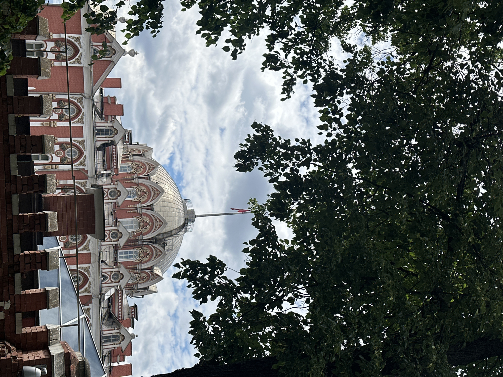 The image shows the upper portion of a historic building with intricate architectural details, including a domed roof and red brickwork with ornate white and gold decorative elements. The building appears to be a grand structure, possibly a palace or a significant public building.
In the foreground and to the right, lush green foliage from a large tree partially obscures the view, creating a natural frame. The leaves are dense, suggesting a warm, possibly summer day. The sky above is a mix of white and grey clouds, indicating it's daytime, though the exact time is not discernible.
A flagpole with a red flag is visible extending from the building's dome area, adding a touch of color against the sky. The overall scene suggests an outdoor setting, with the building likely situated in a park or a leafy urban area. There is no visible text in the image. The perspective is from below, looking upwards, which emphasizes the height and grandeur of the building. The image is oriented vertically.