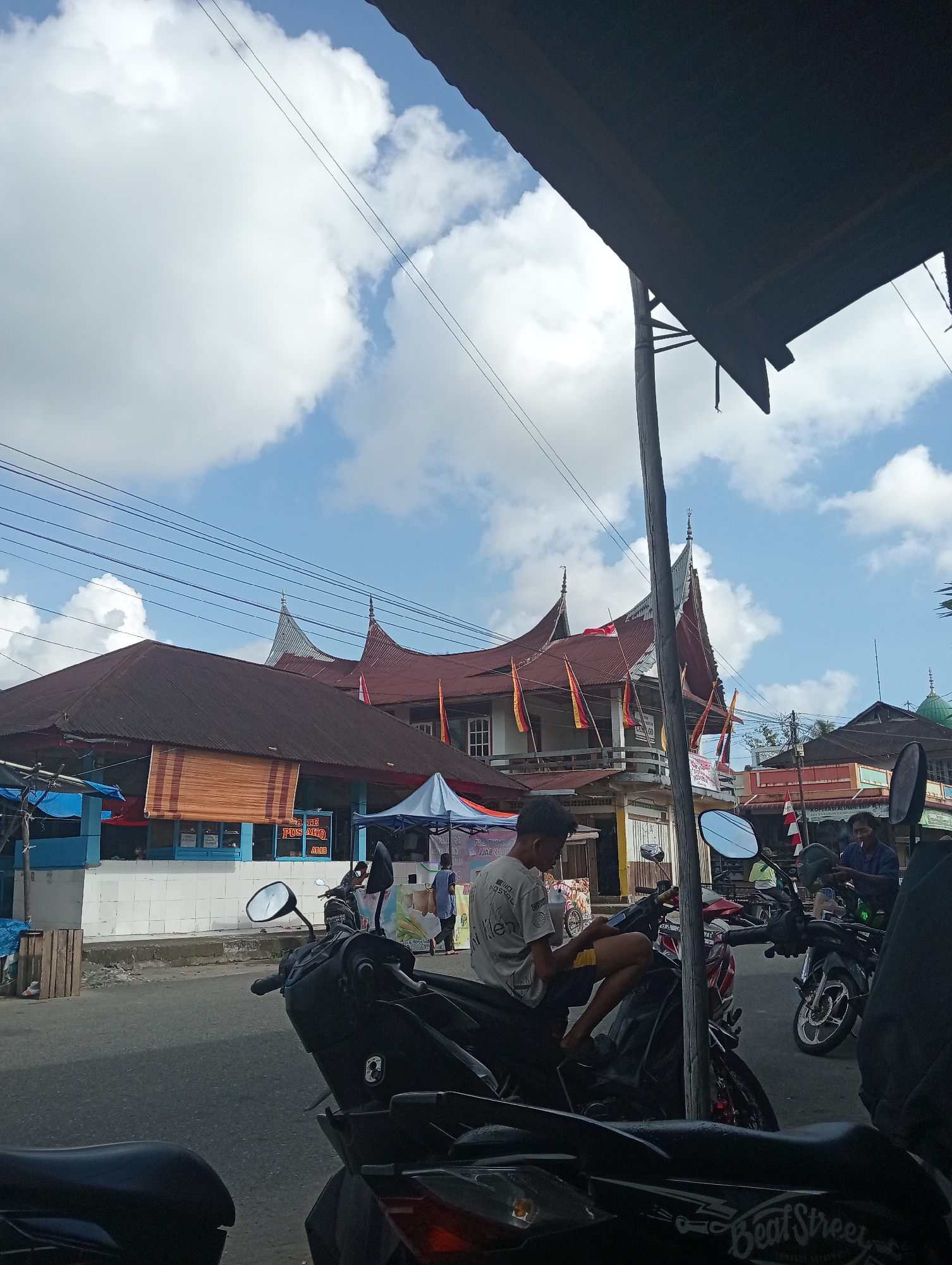 The image depicts a street scene in Pariaman, Indonesia, likely a market area. In the foreground, several motorcycles are parked, with one occupied by a young man sitting on it, looking at his phone. Behind the motorcycles, buildings with traditional Indonesian architecture, characterized by distinctive curved rooflines and red tiles, are visible. Several flags are displayed on the buildings, and some market stalls or awnings are set up in front of them. A few people are walking in the background, and overhead, power lines stretch across the frame against a bright blue sky with scattered white clouds. The overall impression is of a bustling, everyday scene in a Southeast Asian city.