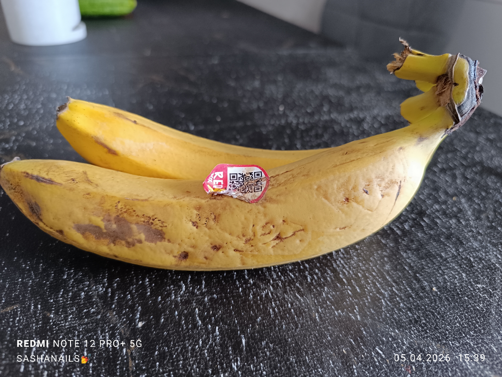 The image shows two ripe bananas lying on a textured, dark gray surface. The banana closest to the viewer has a "REDBOX" sticker with a QR code on it. The bananas are yellow with some brown spots, indicating they are ripening. The background is blurred, but a white container and a green object (possibly a cucumber) are visible. The lower left corner of the image includes the watermark of a Redmi Note 12 Pro+ 5G, and the user's name "SASHANAILS" with a nail polish emoji. The bottom right corner shows the date and time "05.04.2026 15:39".
The location is not specified in the image and it cannot be determined.