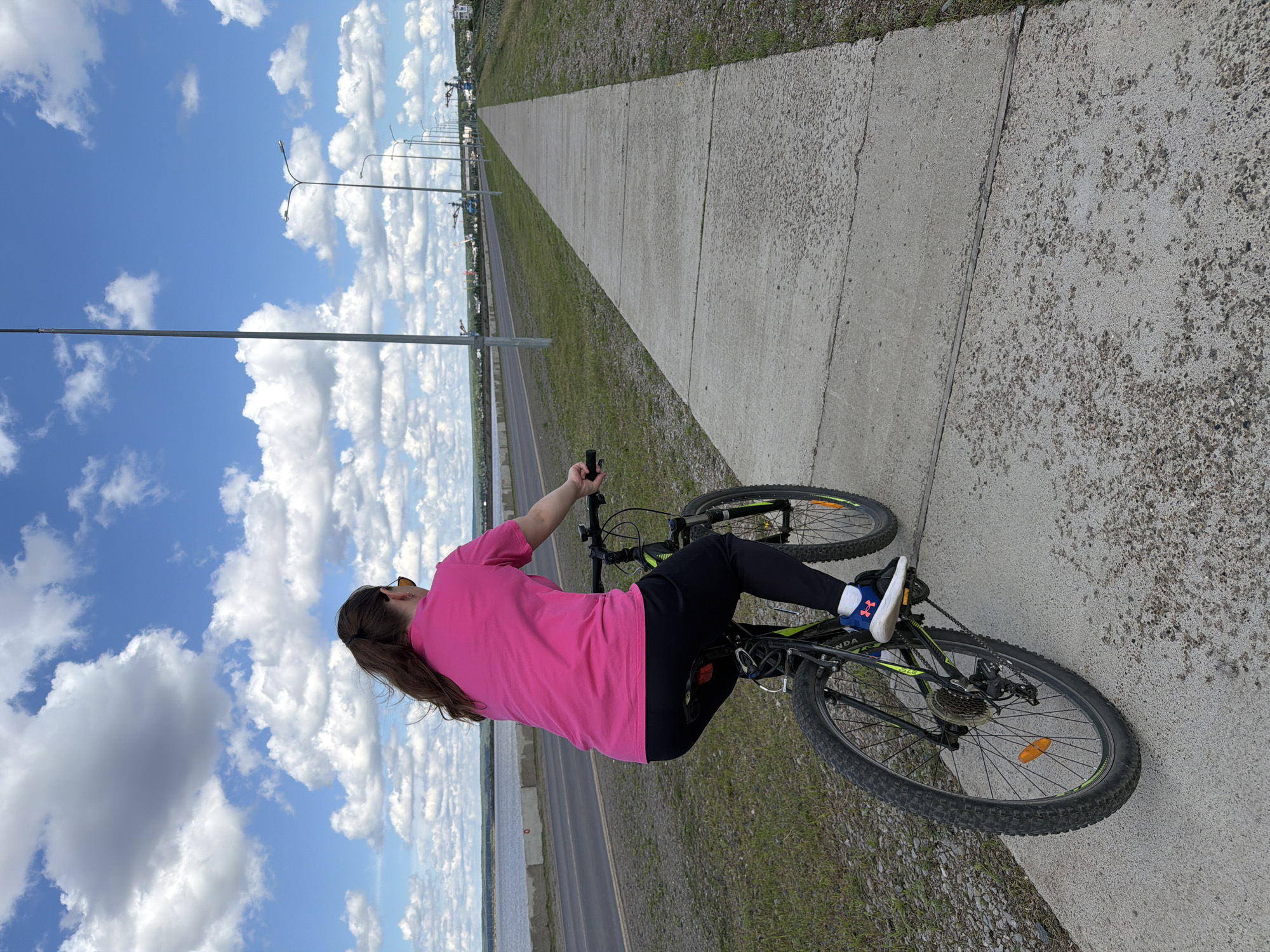 A young woman with brown hair pulled back in a ponytail is riding a black and green mountain bike along a paved pathway. She is wearing a bright pink t-shirt, black pants, and blue and white sneakers. She has sunglasses on. The pathway runs parallel to a road and a body of water, likely the Kama River, with a concrete barrier separating the road from the water. In the background, there are several lampposts, some industrial cranes, and a town or city skyline in the distance. The sky is a vibrant blue with fluffy white clouds. The scene appears to be in the city of Chaikovsky, Russia, possibly along the embankment or a dedicated recreational path.