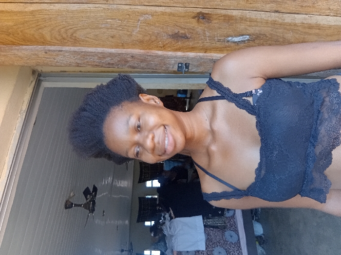 An image taken in Ejule, Nigeria, depicts a young adult female standing in an open doorway. She has dark skin, short, dark, kinky hair styled in an upright fashion, and is smiling, revealing her teeth. She wears a dark blue or black lace crop top with thin straps, beneath which a dark bra is visible. A small earring is present in her left ear. To her right, a weathered wooden doorframe exhibits vertical grain. The background reveals an interior space with a white paneled ceiling equipped with multiple dark-colored ceiling fans, at least two of which are distinct. Furniture draped with patterned fabric, a white garment, and other indistinct objects and figures are visible within the room behind her.