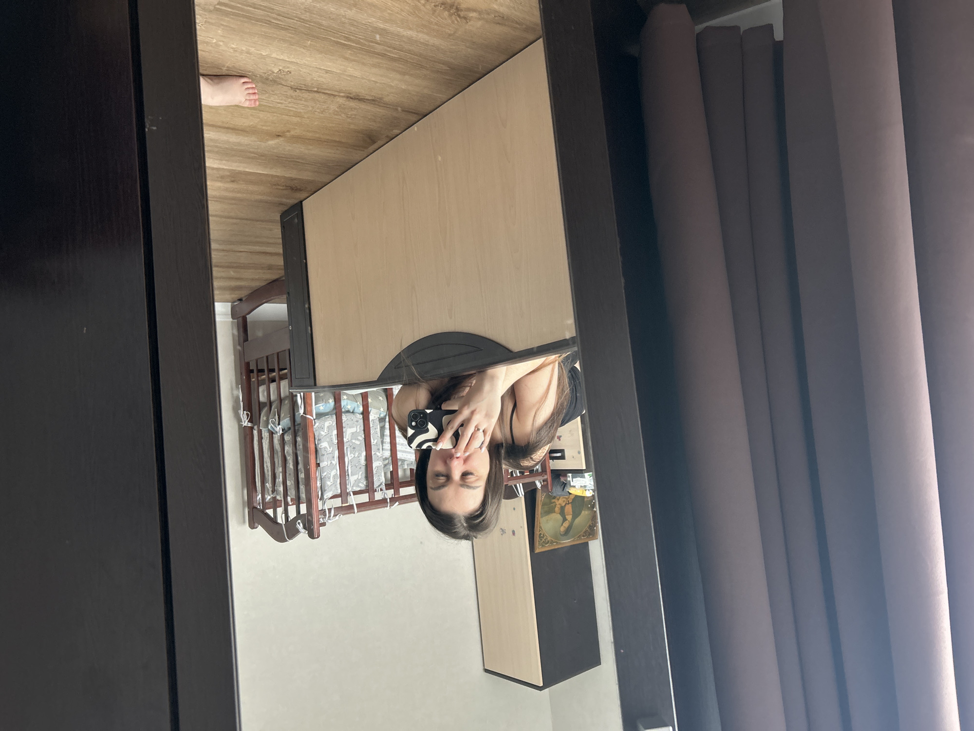 A room is captured in the reflection of a mirror. The walls are light-colored and a crib stands against one of them. A partially visible dresser with a light-colored door and a dark-colored drawer is present. On the wall, there is a framed picture. Brown curtains hang adjacent to the mirror. The mirror reflects a woman holding a phone taking a picture. At the very top of the frame, a bare foot is visible. The location is Chaikovsky, Russia.
