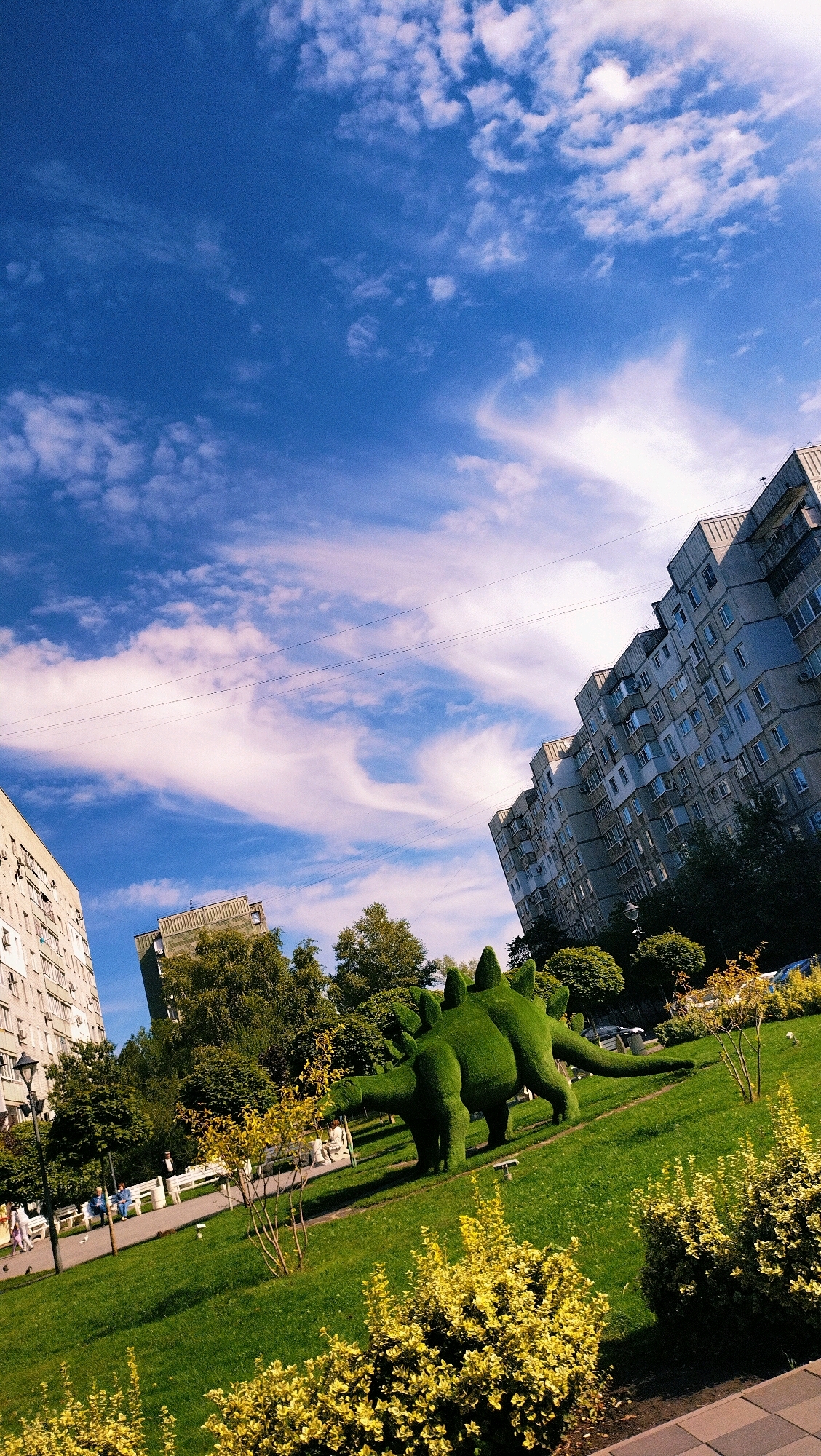 A medium-shot, eye-level outdoor photograph depicts a public park in Horishni Plavni, Ukraine, featuring a topiary dinosaur sculpture. The sculpture, rendered in green shrubbery, resembles a Stegosaurus and is positioned on a well-maintained lawn. In the background, apartment buildings rise under a bright blue sky adorned with wispy white clouds. Several trees, some with yellow foliage, are interspersed throughout the scene, and park benches are visible, occupied by a few individuals. The overall setting suggests a pleasant day in an urban green space.