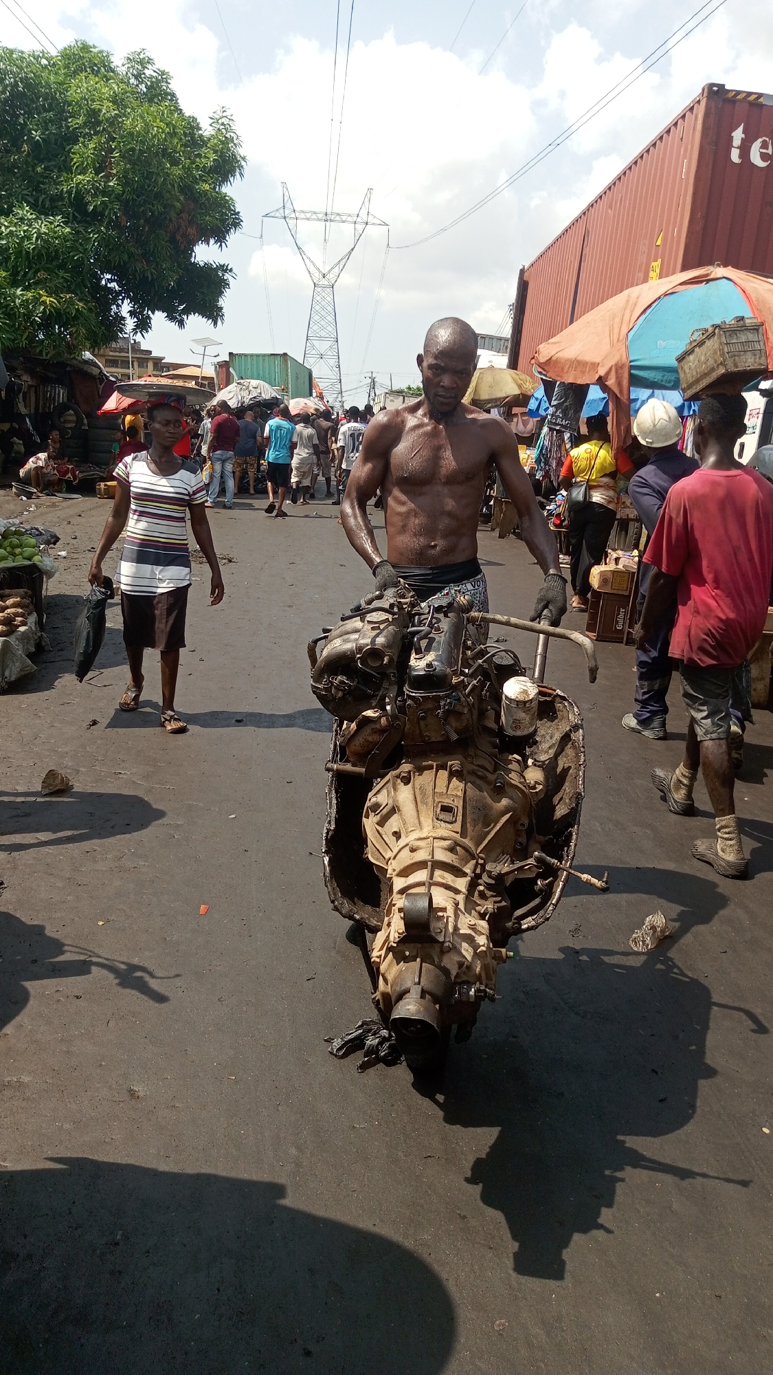This image depicts a bustling street scene in a market area in Onitsha, Nigeria, under bright, harsh midday or early afternoon sunlight, indicated by strong shadows.
The central figure is a shirtless, muscular Black man, glistening with sweat, wearing dark shorts and gloves. He is intensely focused on pushing or pulling a heavy, grimy car engine and transmission assembly, which appears to be mounted on a makeshift frame with handlebars.
Around him, other people navigate the busy street. To his left, a young Black woman in a striped t-shirt and dark skirt walks away, carrying a black bag. To his right, a young Black man in a red t-shirt walks in a similar direction. The background is filled with a lively crowd of people, various market stalls with colorful tarps and umbrellas, and merchandise, including visible green produce on a stall to the left.
In the mid-ground on the right, large red and green shipping containers are prominent. The red container has "te" partially visible on its side. In the far background, a large metal electricity pylon stands tall against a bright, cloudy sky, with power lines extending from it. The ground is a dark, dusty, possibly paved road, scattered with debris. The scene conveys a sense of vigorous activity and commerce in a hot, urban environment.