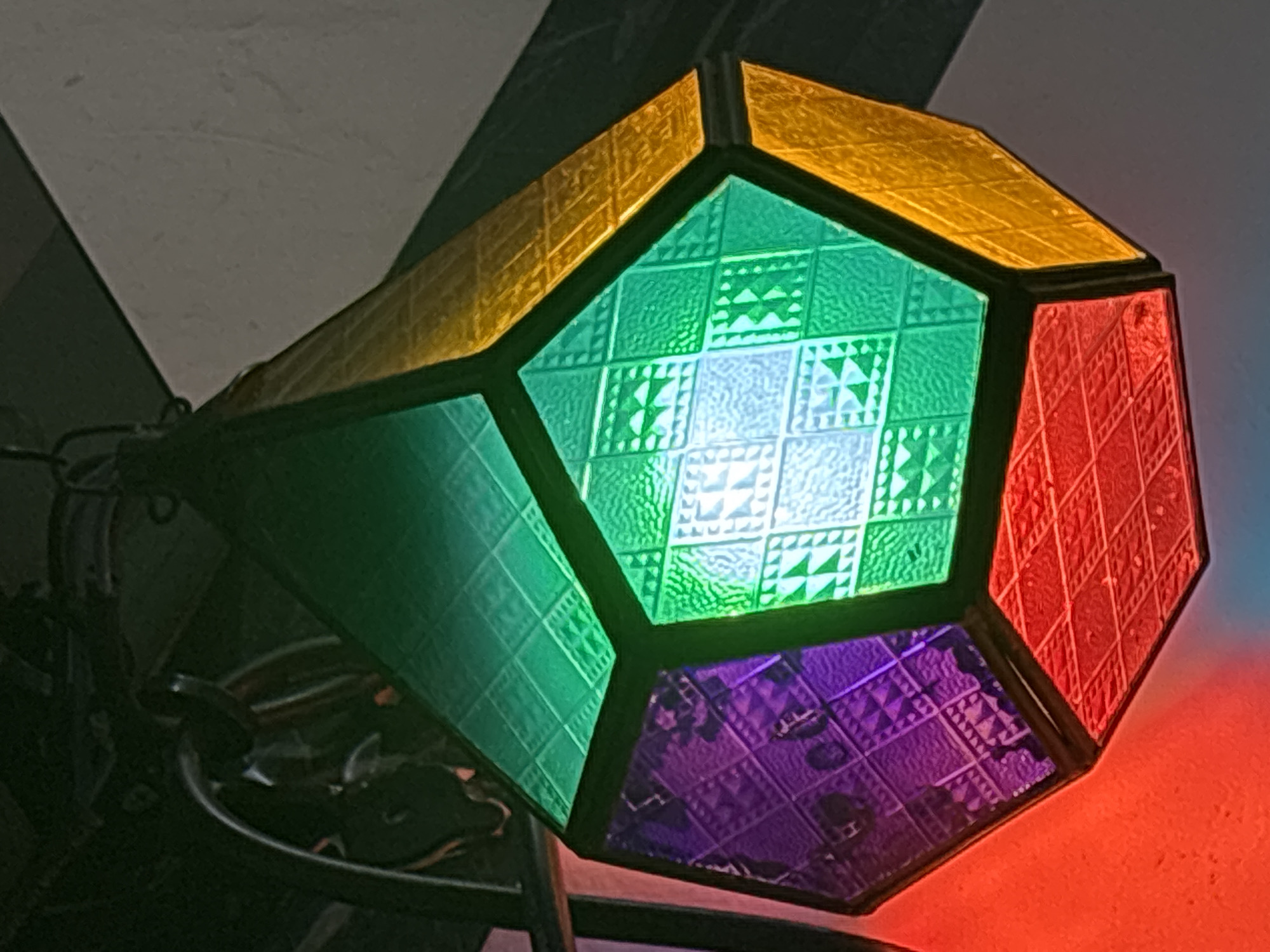 The media file features a vividly colored, geometric lantern or lamp, likely illuminated from within. The lantern is constructed from multiple facets of patterned, translucent material resembling stained glass. The visible panels are bright yellow, emerald green, deep purple, and ruby red. Each panel displays an intricate, textured pattern composed of small squares, triangles, and cross-like shapes, creating a quilted or embossed effect.
A bright light source emanates from inside the lantern, most intensely visible through the central green panel, which has lighter, almost white sections due to the direct illumination. The dark frame holding the colored panels together contrasts sharply with the vibrant hues.
The lantern casts colored light onto the surrounding surfaces, with a noticeable red glow illuminating a textured surface beneath the red panel. The background is dimly lit and largely indistinct, showing dark areas and a light-colored, possibly white or grey, wall or ceiling fragment. The overall impression suggests an indoor setting, likely in low light conditions, emphasizing the decorative and functional aspects of the lantern. There is no visible text in the image. The context for this image is Kuta Selatan, Indonesia.