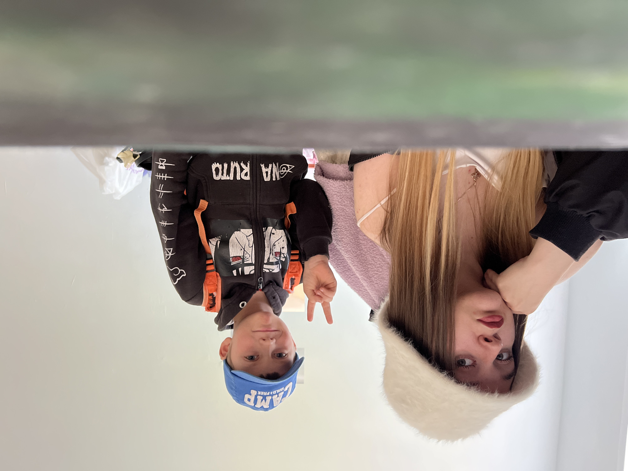 Two individuals are positioned in the lower half of the frame against a pale background. On the left is a young boy wearing a blue cap with the word "CAMP" printed on it, along with a black hoodie featuring orange straps and graphic designs. He is holding up two fingers in a peace sign. To the right of the boy is a young woman with long blonde hair and a white furry hat. She has one hand near her face. Above the subjects is a blurred, indistinct object that obscures the background. The location is unspecified and the image does not give any evidence to identify the city of Pushchino, Russia.