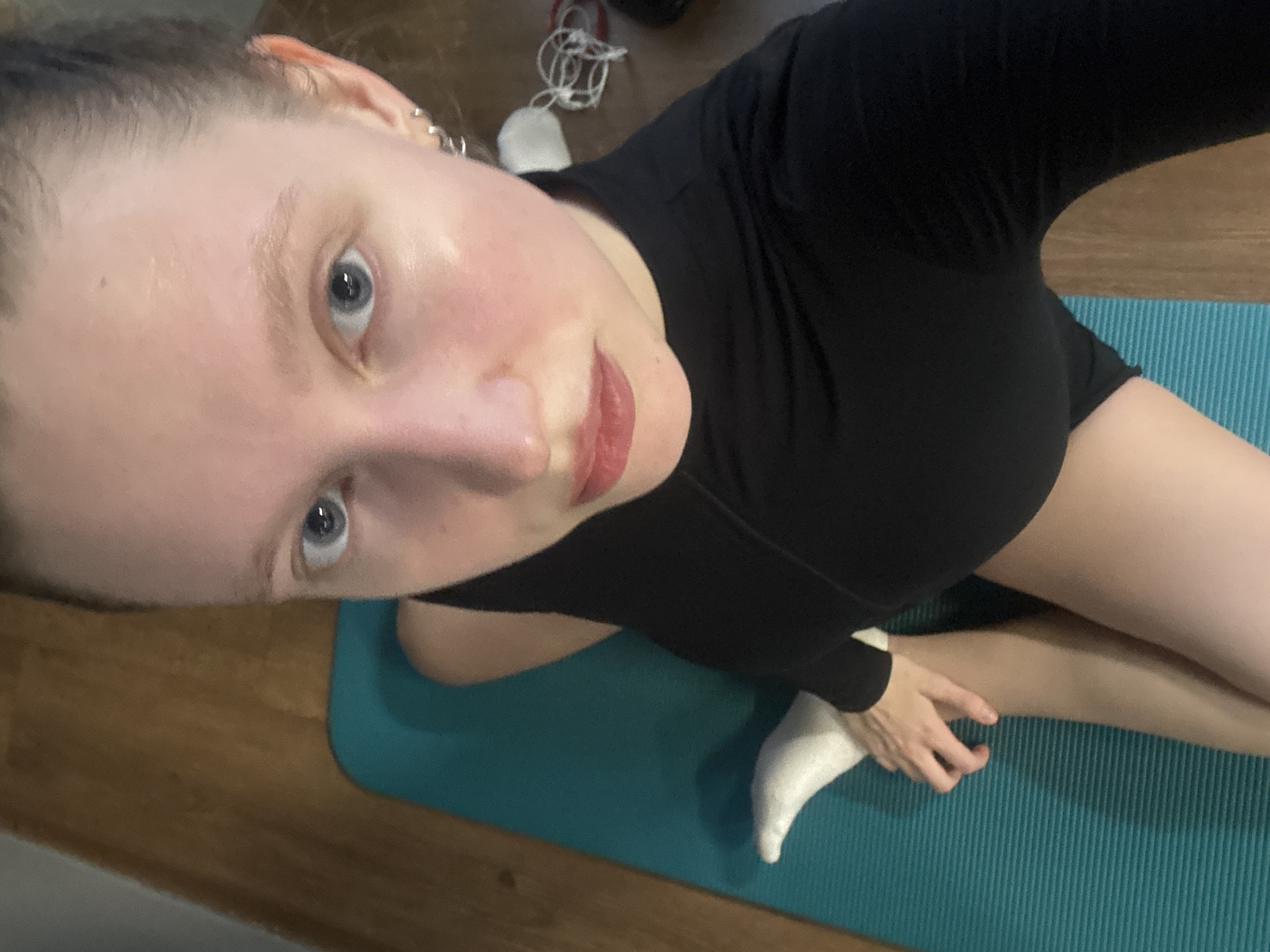 A woman is depicted in a high-angle, close-up indoor selfie. She has fair skin, light blue eyes, and light-colored hair pulled back from her face. Small silver-toned hoop earrings are visible in both ears. She is wearing a black long-sleeved athletic top with a mock neck and black athletic shorts, with a white sock on her left foot. Her left hand is resting on her right thigh. The woman is seated or kneeling on a ribbed, teal-colored yoga mat, which is placed on a light brown wooden floor. A light-colored wall is partially visible in the upper left background. In the blurred background to the right, a white fabric object and tangled white wires are discernable. The specific city or country of Izhevsk, Russia, or any other location, cannot be identified from the visual information presented in this image.