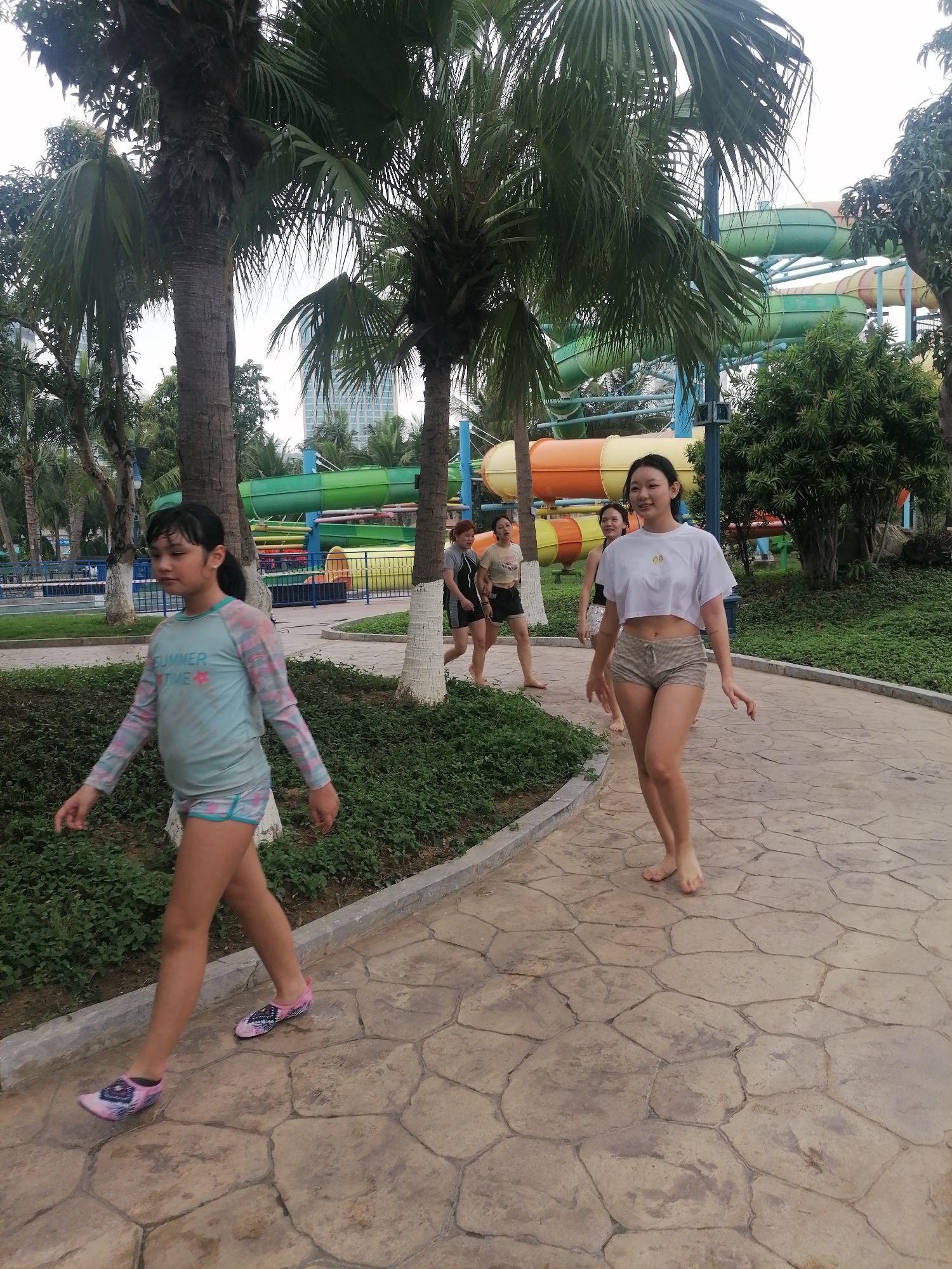 A group of people, predominantly young women and girls, are captured walking along a paved pathway within what appears to be a tropical water park or resort in Bai Chay Ward, Vietnam. The scene is brightly lit, suggesting daytime.
In the foreground, a young girl with dark hair walks towards the left side of the frame. She wears a light blue long-sleeved swim shirt featuring the text "SUMMER TIME" and pink star motifs, paired with matching patterned shorts and pink and black water shoes. Behind her, a young woman with dark hair smiles warmly as she walks barefoot towards the viewer. She is dressed in a white cropped t-shirt, which has two small yellow fruit emojis near the collar, and light brown patterned shorts. Several other individuals, some also barefoot and wearing casual or swimwear, can be seen further back, walking along the path.
The pathway consists of irregularly shaped, light-colored paving stones and is lined with green bushes and ground cover. Numerous tall palm trees are scattered throughout the environment, contributing to the tropical atmosphere. In the background, colorful water slides in green, orange, and yellow hues are prominent, winding through the lush greenery, confirming the water park setting. The overall impression is one of a relaxed and enjoyable outing.