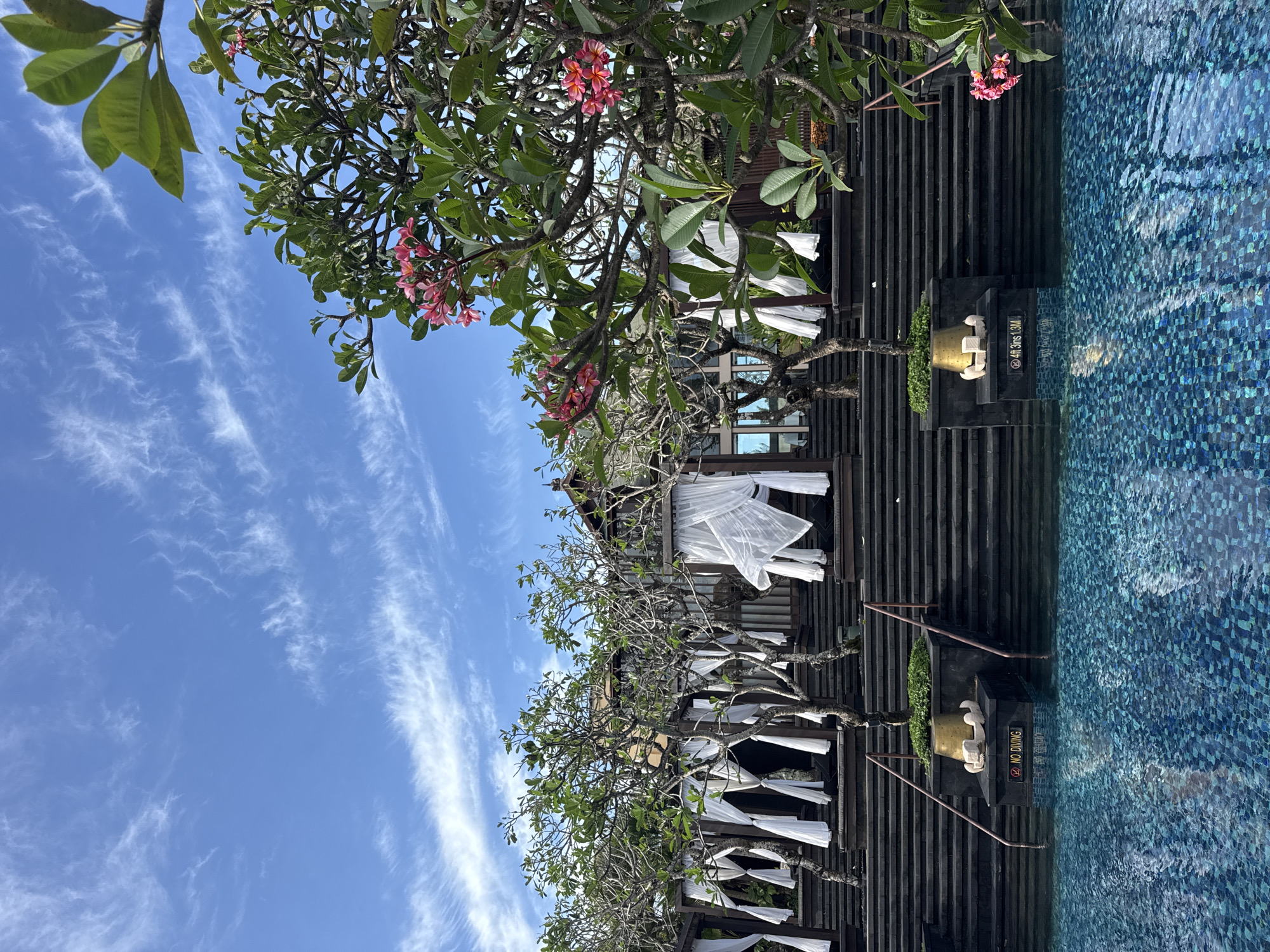This is a wide-angle, eye-level outdoor shot of a resort pool area in Kuta Selatan, Indonesia. The water in the foreground is a deep blue with a tiled mosaic pattern. In the mid-ground, a wide, dark, stepped structure rises from the pool's edge, leading to a building. White curtains are draped on structures along the sides of the steps, suggesting cabanas or seating areas. There are trees with green leaves and pink flowers scattered throughout the scene. The sky above is a bright blue with wispy white clouds. Two signs are visible near the pool's edge: one reads "NO DIVING" and the other indicates "4FT 3INS 1.3M".