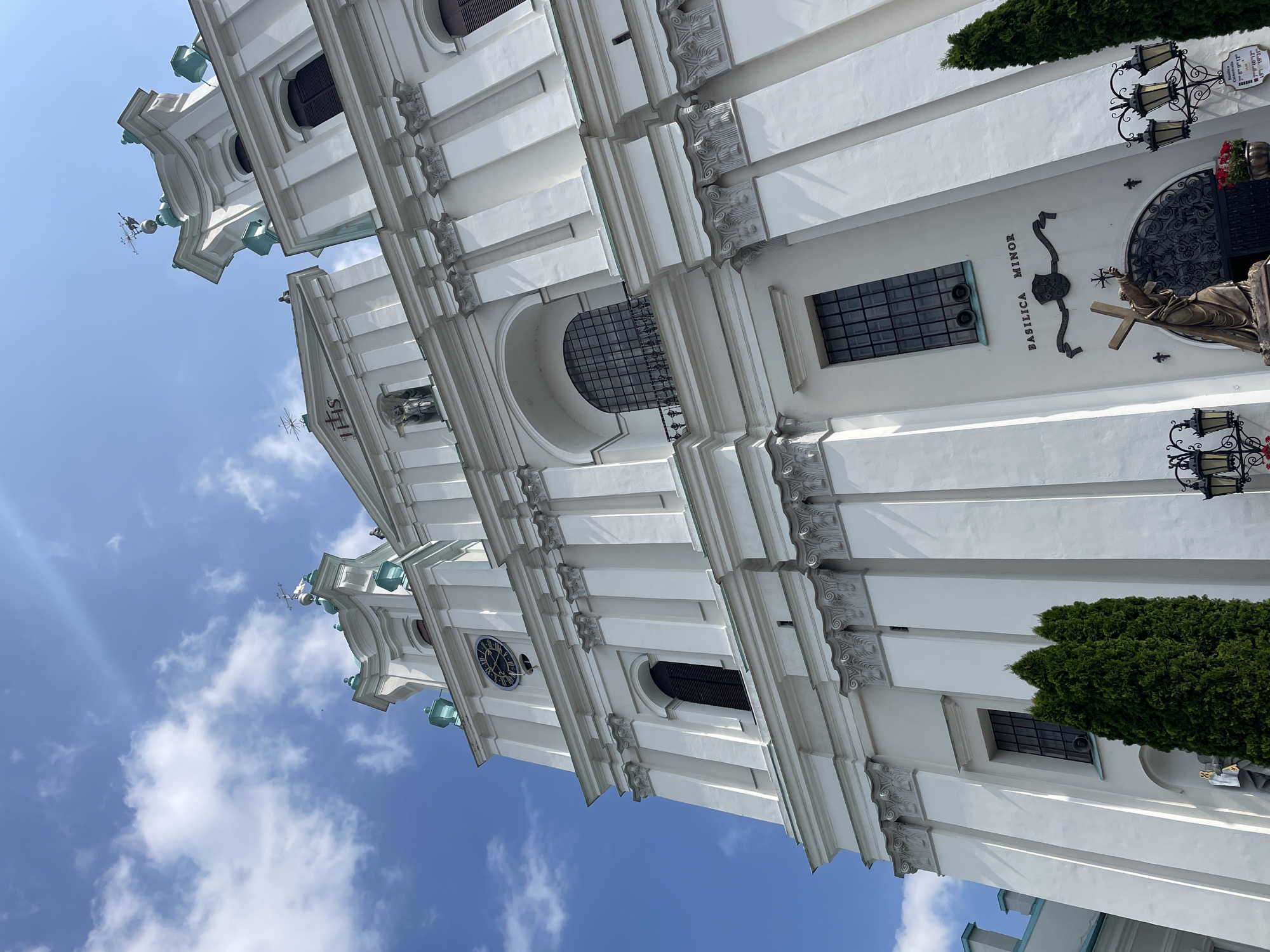 This is a low-angle, outdoor, daytime photograph of the exterior of the Saint Francis Xavier Cathedral in Hrodna, Belarus. The white Baroque facade of the cathedral is prominently featured, with its tall towers, ornate architectural details, and a large clock on one of the towers. The sky above is blue with scattered white clouds, and sunlight casts shadows on the building. In the foreground, there are evergreen trees and a statue of a man holding a cross. The text "BASILICA MINOR" is visible above the entrance. The overall impression is one of grandeur and religious significance.