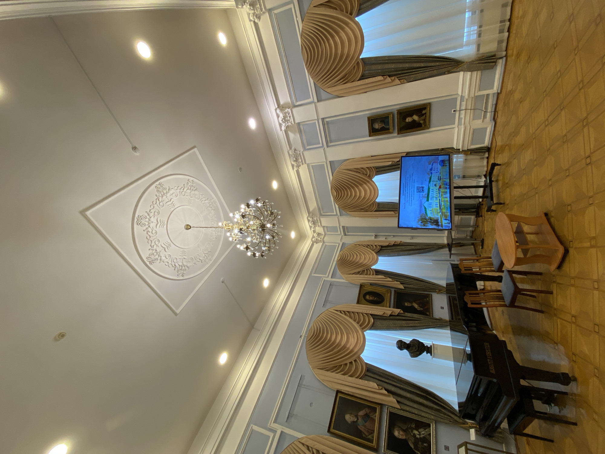 The image depicts an interior scene of a room that appears to be a museum or a grand hall, possibly located in Petrozavodsk, Russia. The room features a high ceiling with ornate molding and a chandelier. There are several framed portraits hanging on the pale blue walls, some with elaborate drapes over arched windows.
In the foreground, a black grand piano with the inscription "AUGUST FÖRSTER" is visible. To the right of the piano, there are two chairs and a small wooden table. Further to the right, a large flat-screen television on a stand displays an image of a building and text, likely part of an exhibition or presentation. A bust of a man is positioned on a pedestal behind the piano. The floor is made of polished wood with a patterned inlay. The lighting appears to be a combination of recessed ceiling lights and the chandelier. There are no people visible in the frame.