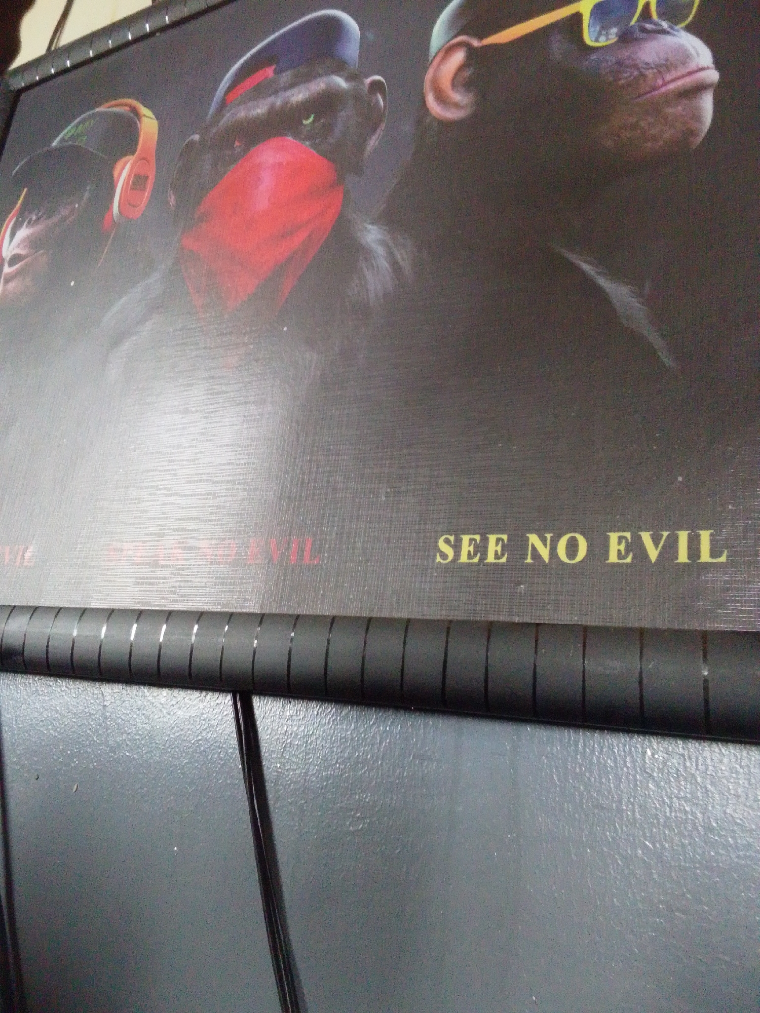 The image shows a close-up of a poster featuring three chimpanzees. The chimpanzee on the left wears orange headphones and a black cap. The middle chimpanzee wears a red bandana over its mouth and a black cap. The chimpanzee on the right wears yellow sunglasses. The text "SEE NO EVIL" is visible in yellow lettering on the poster. Below the poster is a black bar with reflective vertical lines. The background is a textured gray surface, possibly a wall or a desk. The image is taken from a slightly angled perspective, and the lighting is not uniform. There are no visible indicators of the city or location within the image.