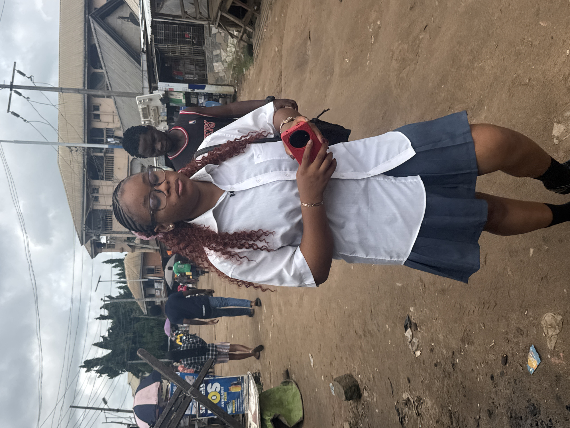 A young woman with reddish-brown curly hair and glasses, wearing a white collared shirt and a grey pleated skirt, walks on a dirt road in Ihiagwa, Nigeria. She is holding a red object in her hands and has a black bag slung over her shoulder. Behind her, a young man wearing a black tank top and jeans walks in the same direction. The environment is a bustling street with buildings, market stalls, and overhead power lines. The ground is unpaved and appears dusty, with scattered debris. Other people and parked vehicles are visible in the background, suggesting a busy, everyday scene. The sky is overcast.